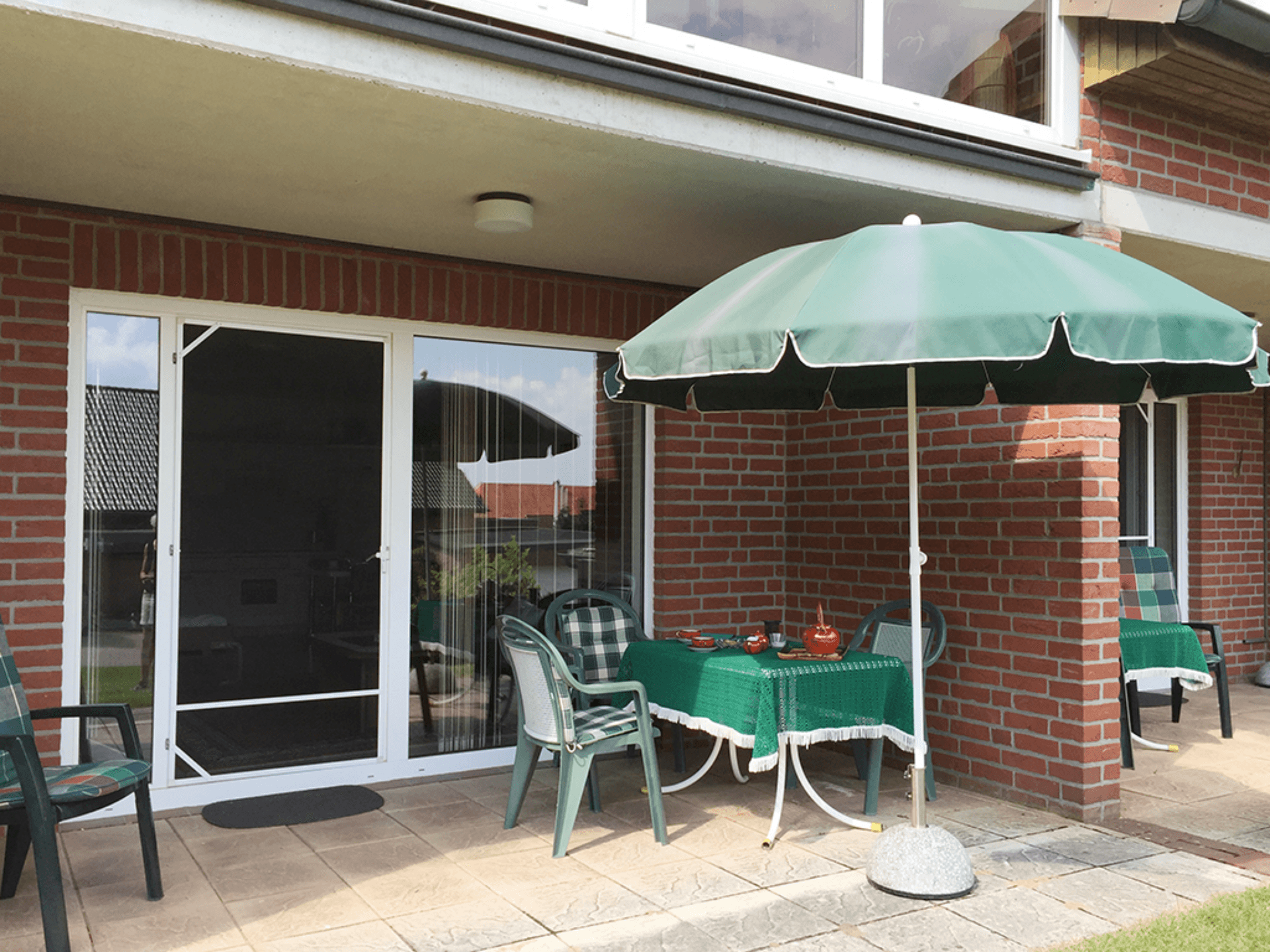 Sonnige Terrasse am Backsteinhaus mit gedecktem Tisch, Stühlen und grünem Sonnenschirm.