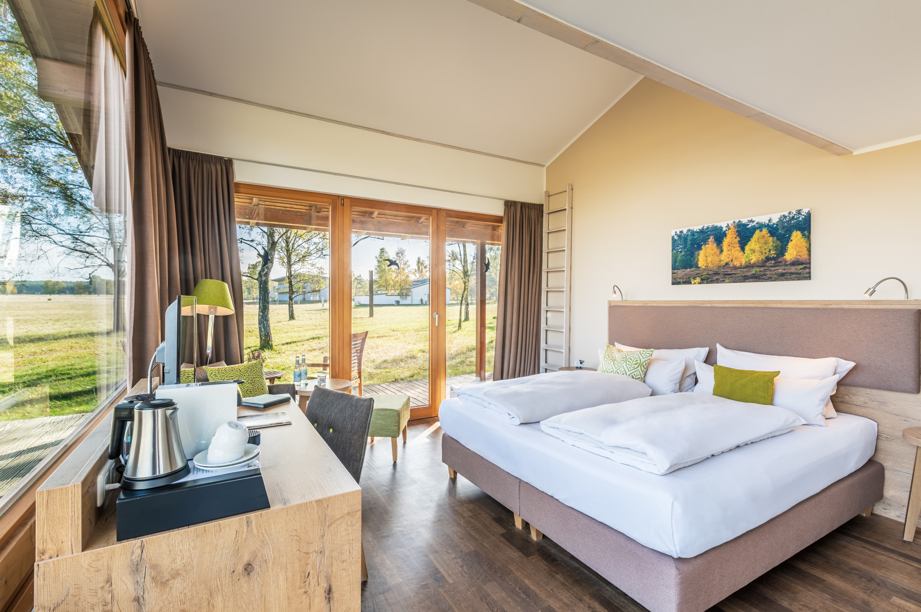 Innenbereich des Hotelzimmers. Ein gemachtes Bett, ein Schreibtisch, eine große Fensterfront mit einem Ausblick in die Weite der Natur.