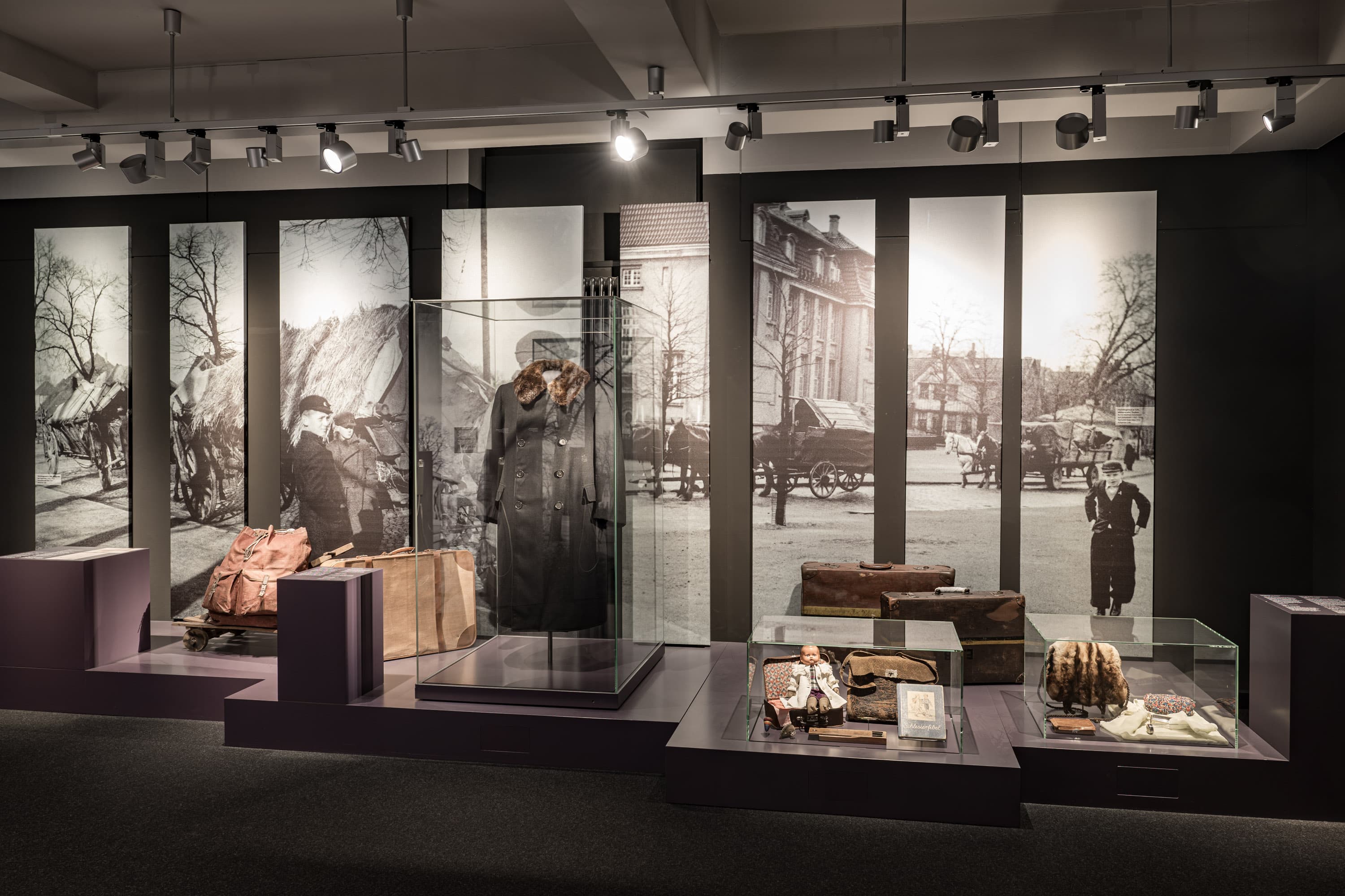 Ausstellungsansicht zur Fluchtgeschichte mit Koffern, Mantel und großen Schwarz-Weiß-Fotografien.