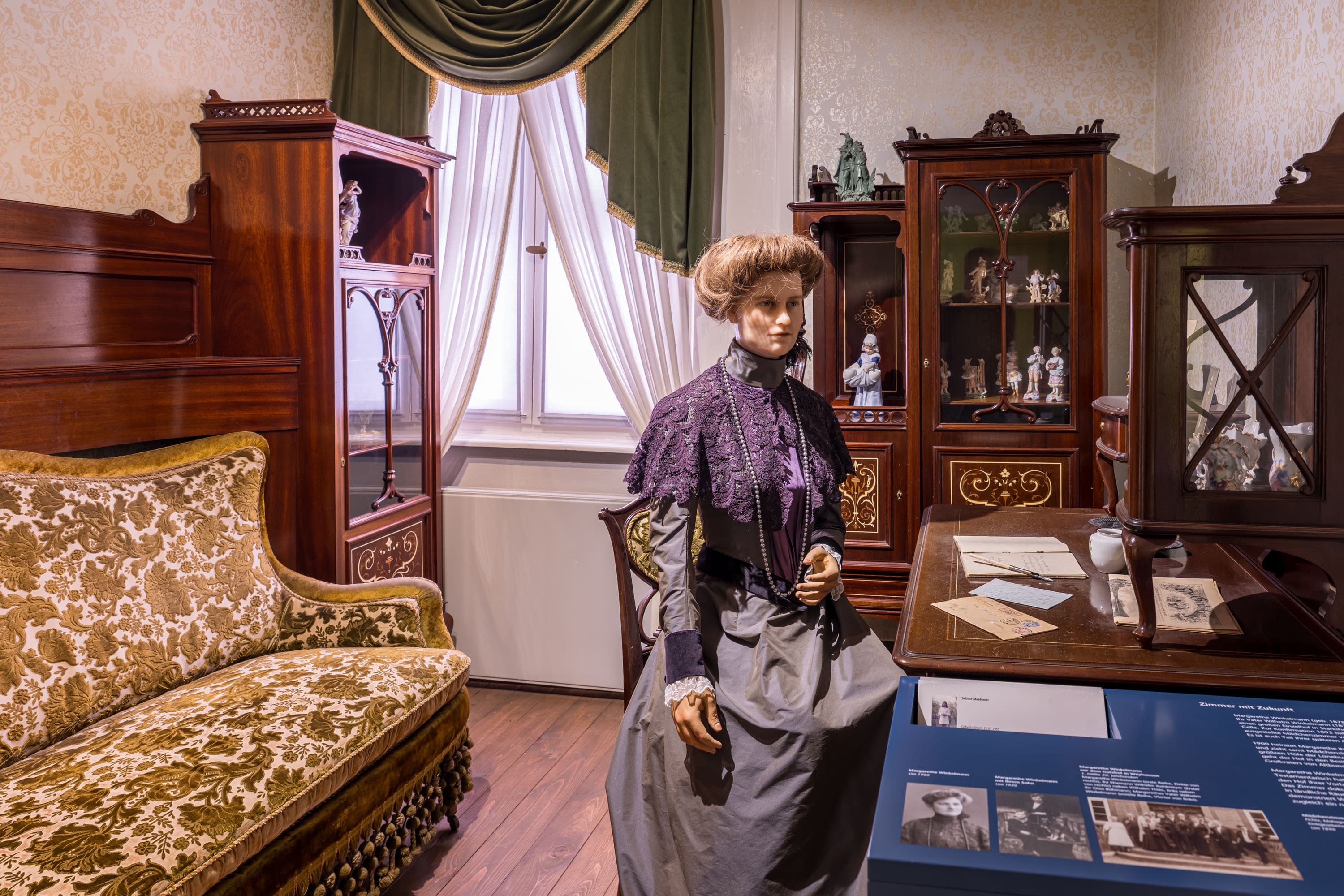 Eine weibliche Figur in historischer Kleidung in einer nachgestellten bürgerlichen Wohnstube um 1900.