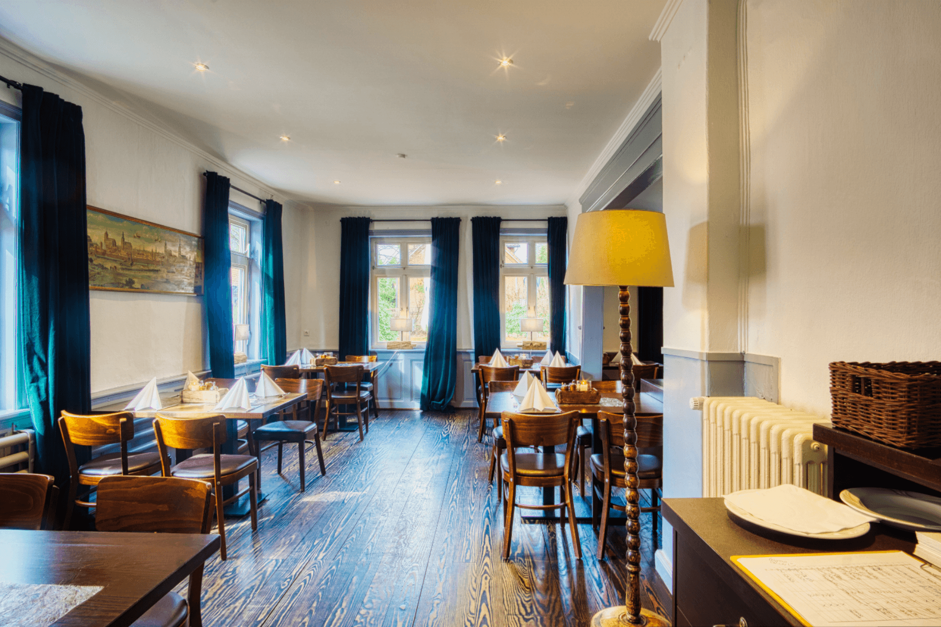 Heller Gastraum mit Holzfußboden, eingedeckten Holztischen und langen dunklen Vorhängen.