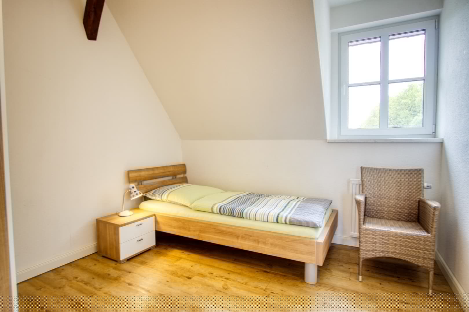 Einzelbett in einem Schlafzimmer unterm Dach mit Holzboden, Nachttisch und einem Korbsessel.