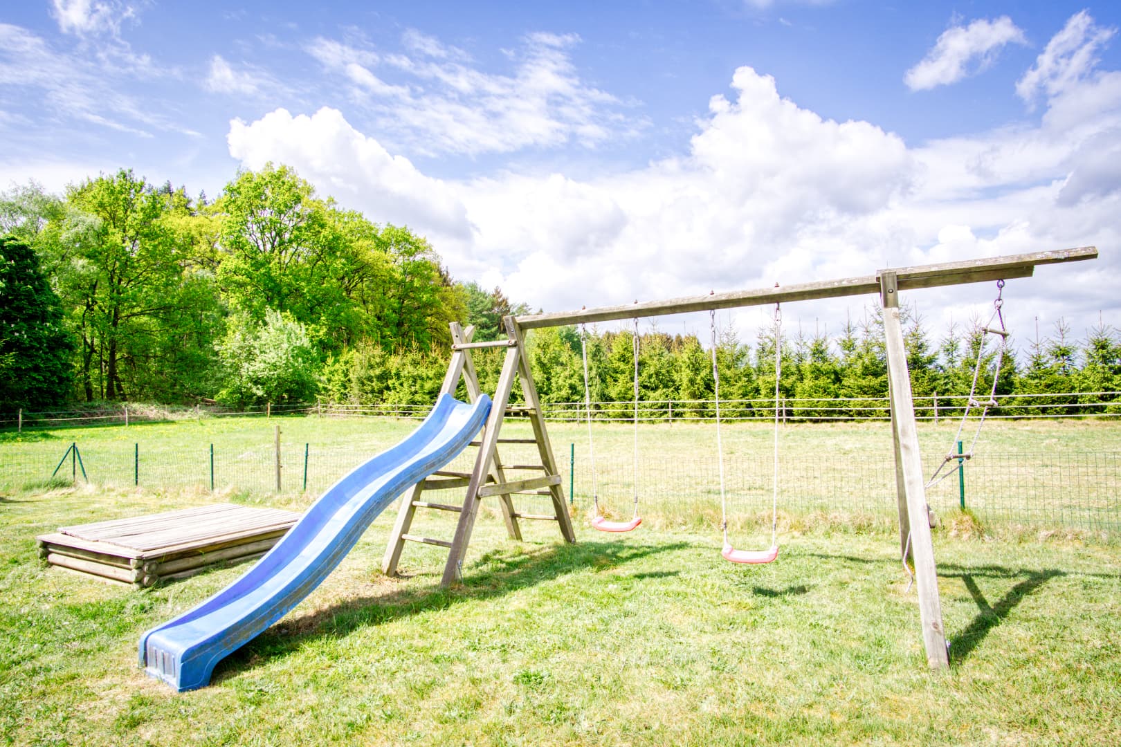 Holzspielgerüst mit blauer Rutsche und zwei roten Schaukeln auf einer Wiese am Waldrand.