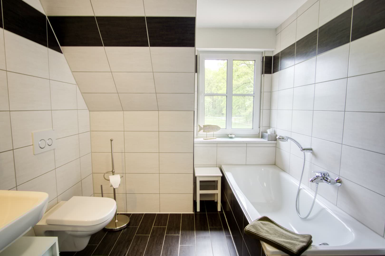 Hell gefliestes Badezimmer mit Badewanne, WC, dunklem Boden und einem Fenster mit Waldblick.