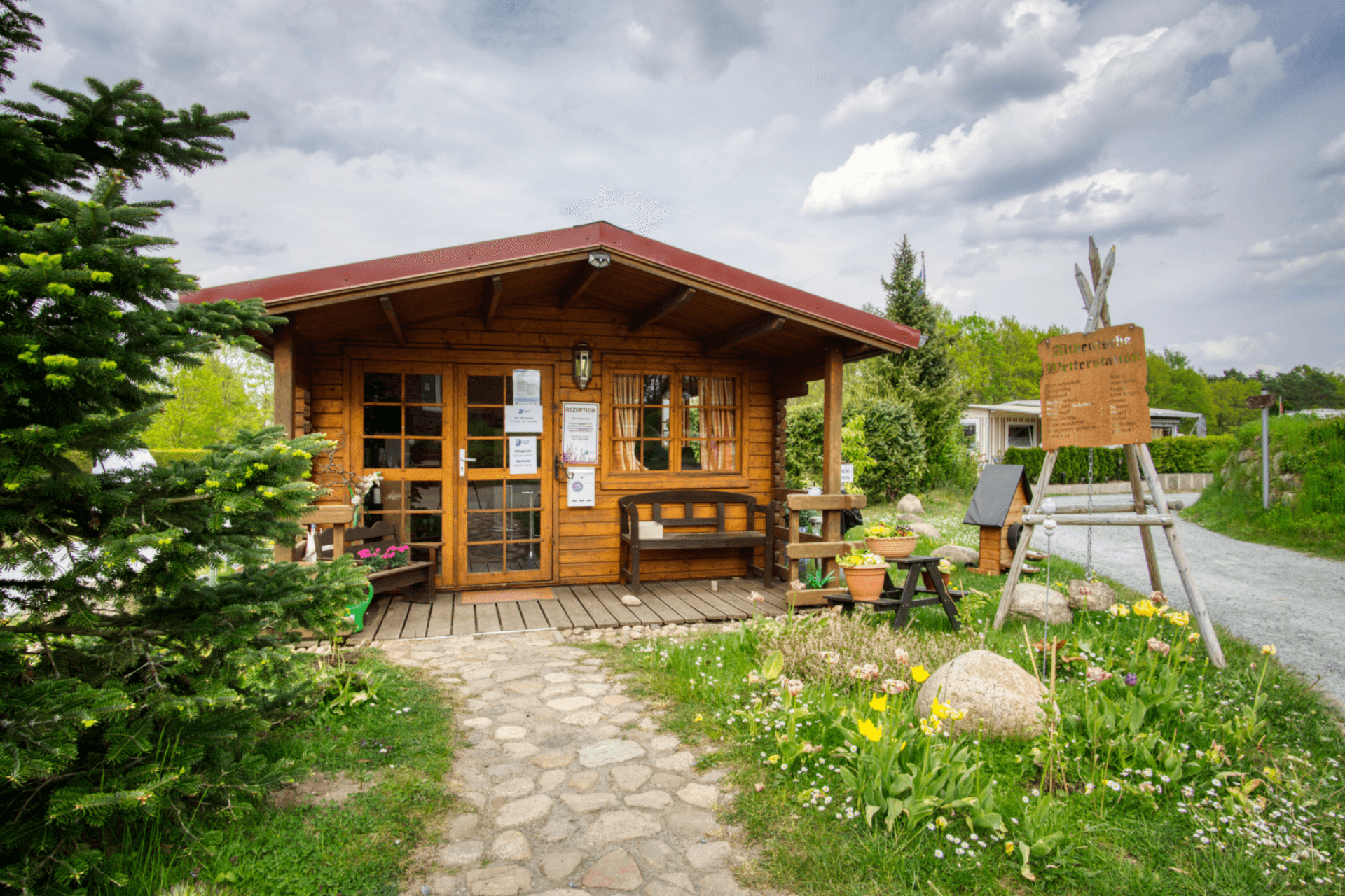 Kleines Holzhaus mit rotem Dach und Blumenschmuck dient als Rezeption des Campingplatzes Lopautal. Ein gepflasterter Weg führt zum Eingang, umgeben von grüner Natur und bunten Blumen, die eine einladende Atmosphäre schaffen.
