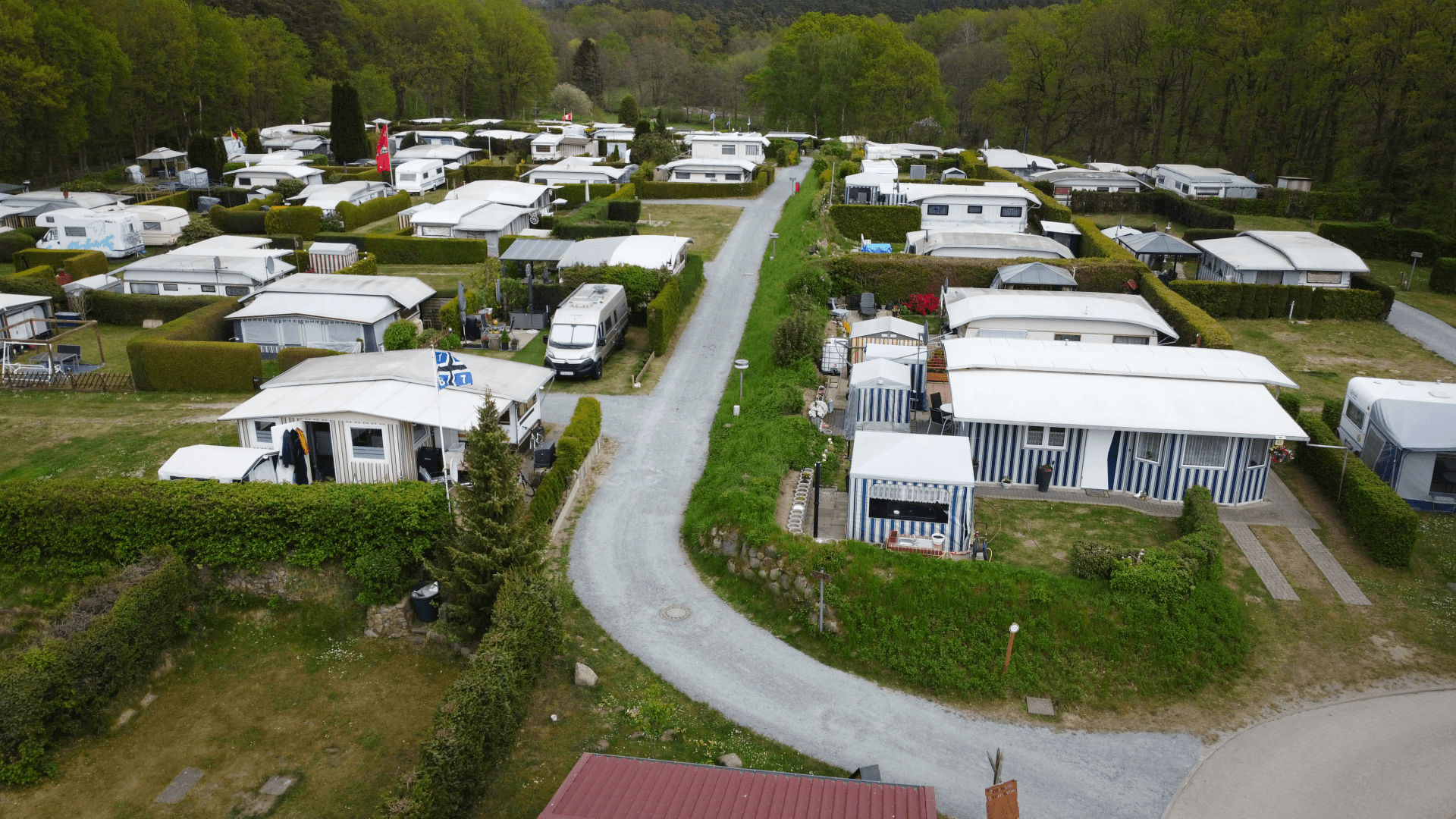 Blick über den Campingplatz Lopautal mit zahlreichen Wohnwagen und Mobilheimen, getrennt durch gepflegte Hecken und Wege, umgeben von dichten Laubbäumen am Waldrand unter leicht bewölktem Himmel.