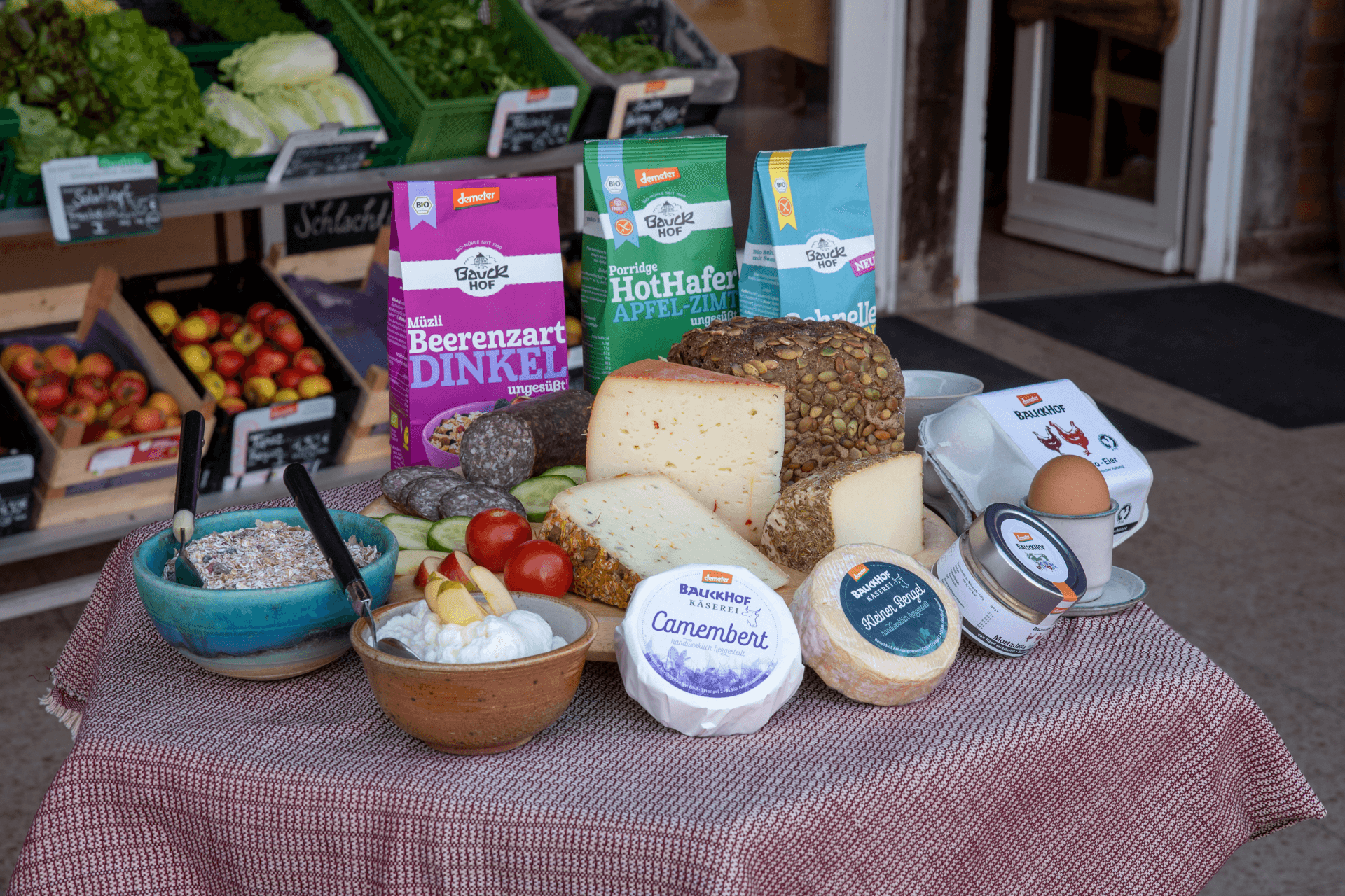 Das Bild zeigt eine liebevoll arrangierte Auswahl an biologischen Backwaren, verschiedenen Käsesorten und Müsli-Schalen, die appetitlich auf einem Tisch drapiert sind. Im Hintergrund sind frisches Bio-Gemüse und Obst zu sehen, die die Vielfalt und Frische des Sortiments unterstreichen.