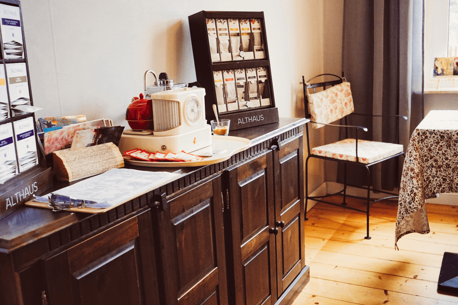 Holzschrank mit Teeauswahl, Retroradio und Speisekarte neben einem Tisch mit floraler Decke.