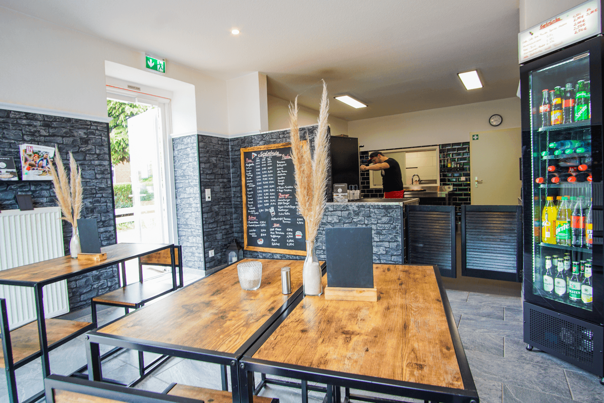 Innenraum eines modernen Restaurants mit Holz-Metall-Tischen, schwarzer Steinoptik an den Wänden und einer gut sichtbaren Speisetafel. Im Hintergrund bereitet eine Person Speisen in der offenen Küche zu, rechts steht ein beleuchteter Kühlschrank mit Getränken.