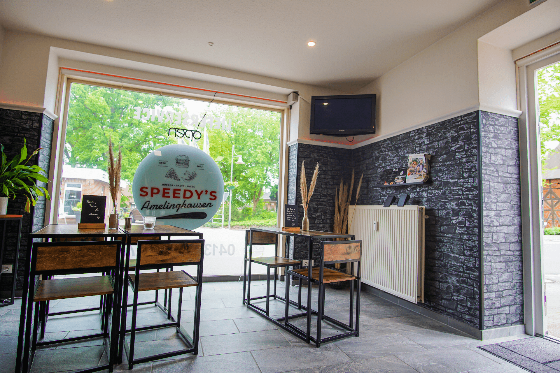 Heller Innenraum des Restaurants Speedy’s Amelinghausen mit großen Fenstern, modernem Mobiliar aus Holz und Metall sowie dunklen Steinwänden. Auf dem Fenster ist das Restaurantlogo zu sehen, draußen grünes Laub. Ein Fernseher hängt an der Wand, Dekoelemente sorgen für ein modernes Ambiente.