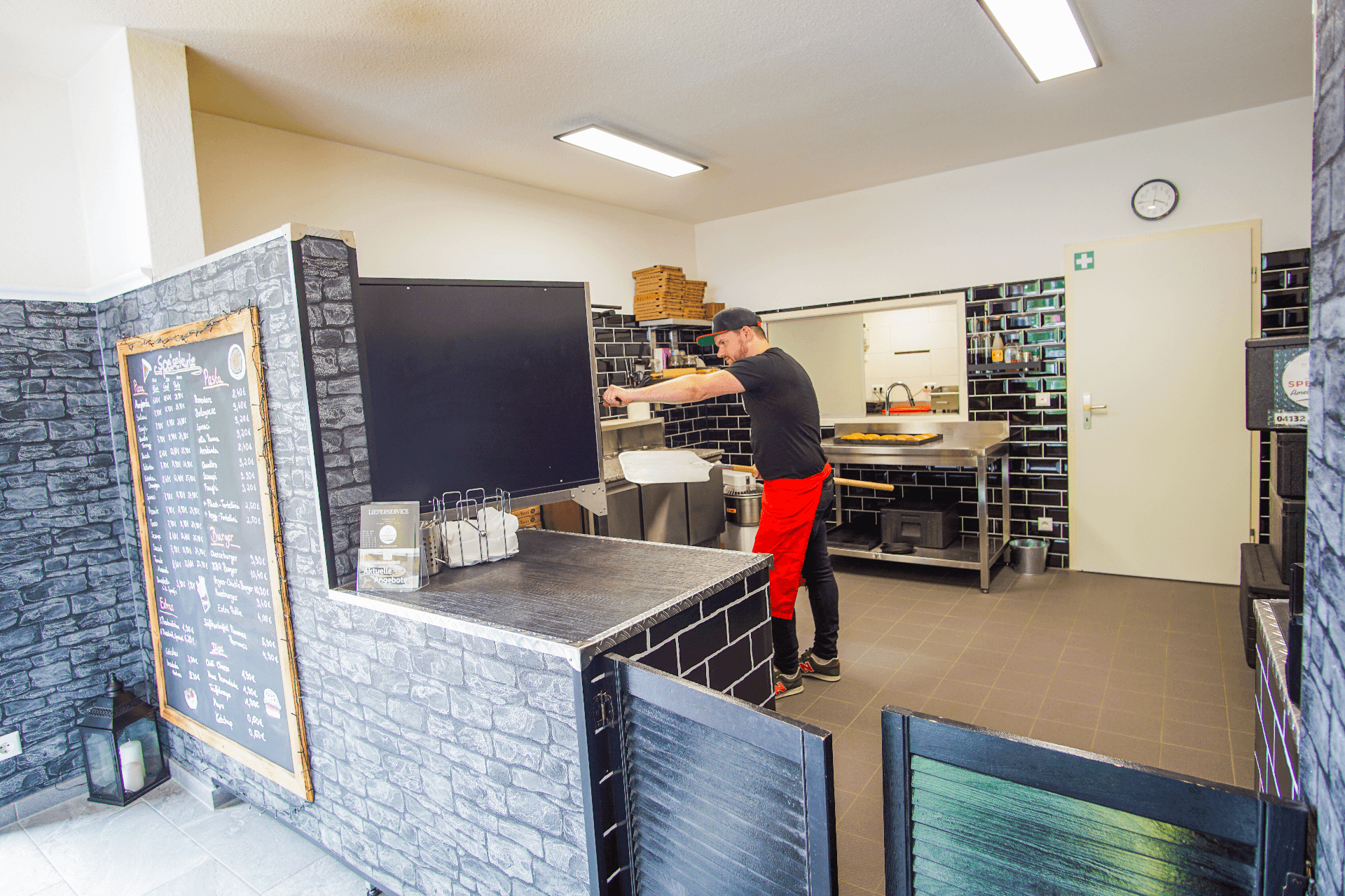 Ein Mitarbeiter formt Teig in einer modernen Pizzaküche mit dunklen Fliesen und heller Beleuchtung. Im Vordergrund steht eine Theke mit Speisekarte, im Hintergrund liegen Pizzaschachteln und Küchenutensilien bereit. Die Umgebung wirkt sauber und barrierefrei gestaltet.