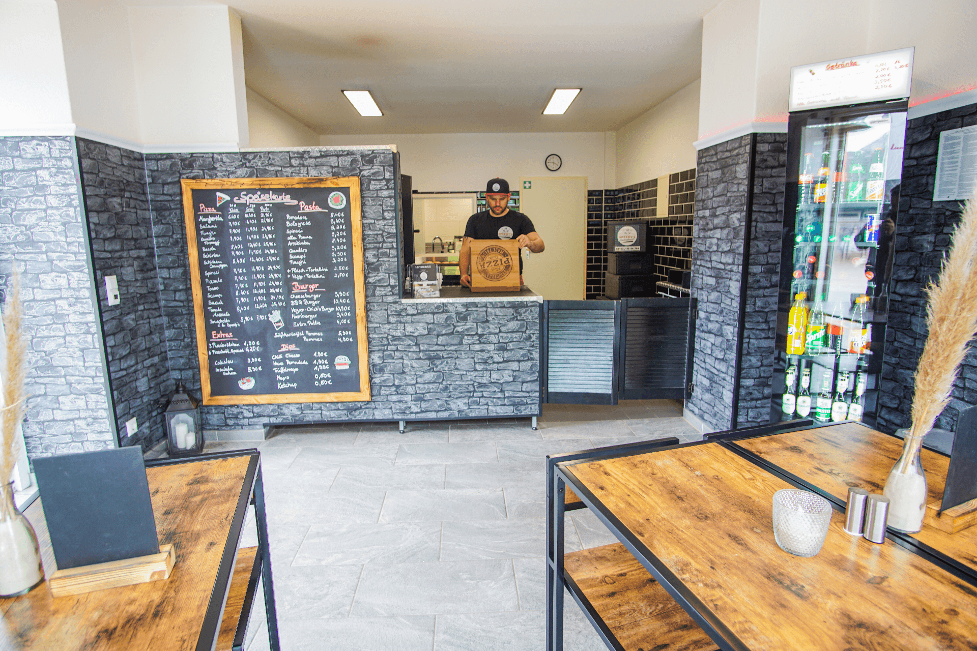 Heller Gastraum mit modernen Holztischen, einer Getränkekühlvitrine und einer großen Menütafel an der Wand. Hinter der Theke steht ein Mitarbeiter mit einer Pizzaschachtel. Die Einrichtung wirkt klar strukturiert, barrierefrei zugänglich und auf Selbstbedienung ausgelegt.