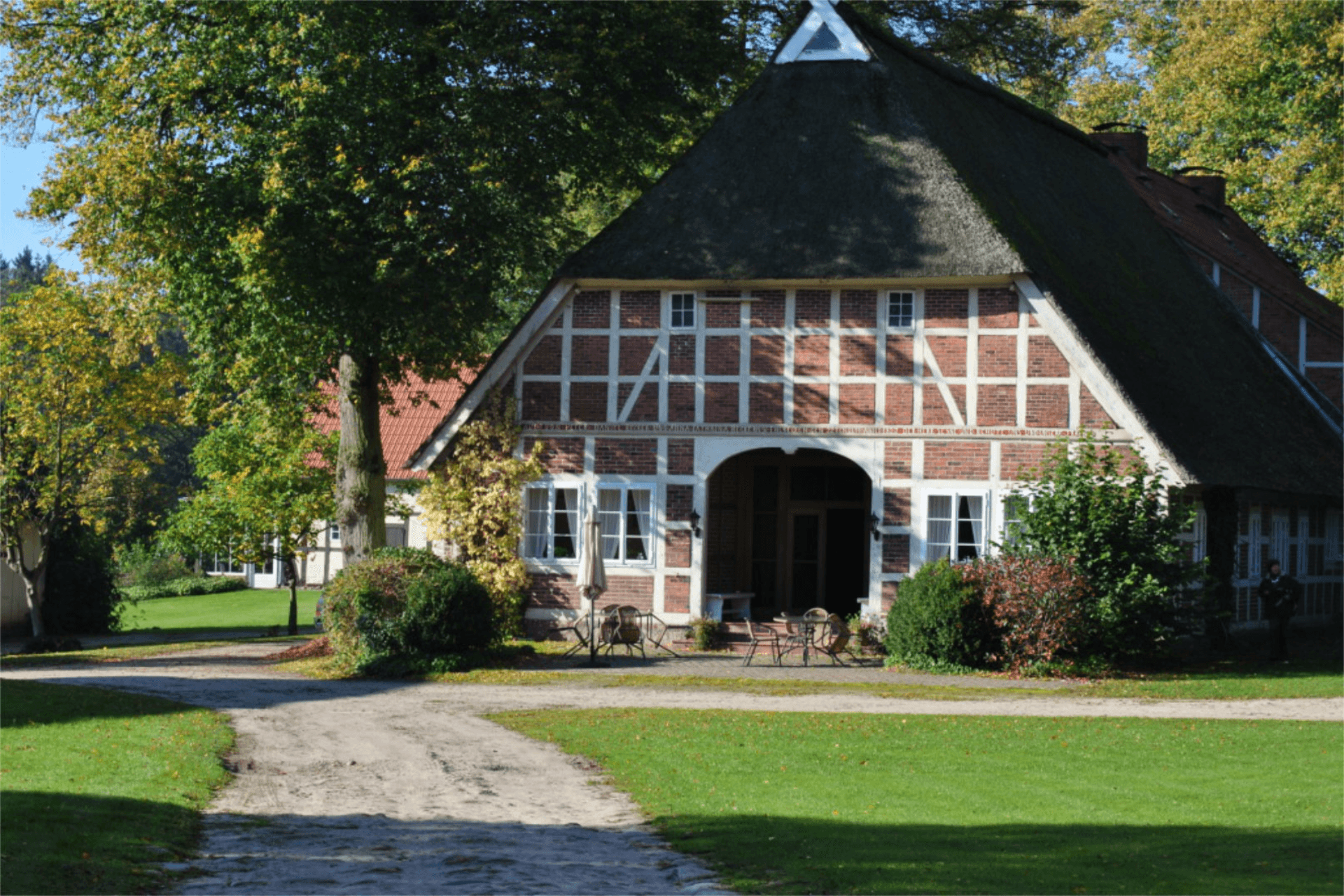 Historisches Bauernhaus mit rotem Ziegelfachwerk und Reetdach, umgeben von grüner Wiese und alten Bäumen. Vor dem Eingang stehen Gartenmöbel im Sonnenschein, der Hofweg aus Sand führt durch eine ruhige, naturreiche Landschaft – ein Ort zum Entspannen und Entdecken.