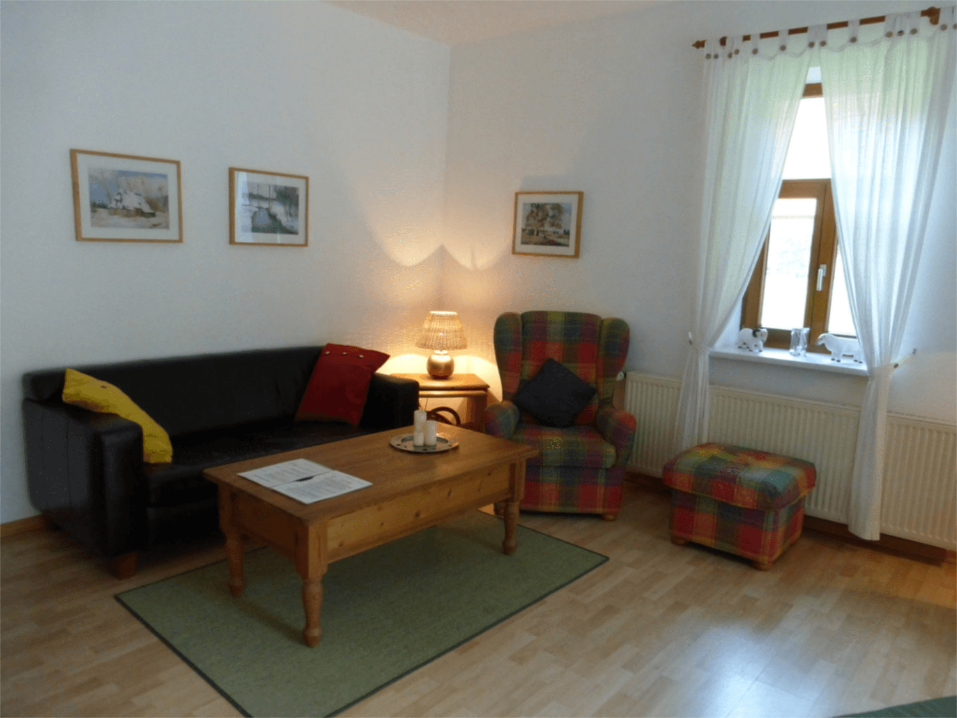 Helles Wohnzimmer mit Holzfußboden, schwarzem Ledersofa und farbenfrohem Sessel im Karomuster. Ein Holztisch mit Kerze steht auf einem Teppich in der Mitte. Neben dem Fenster mit weißen Vorhängen sorgt eine Tischlampe für warmes Licht und schafft eine einladende Atmosphäre zum Entspannen.