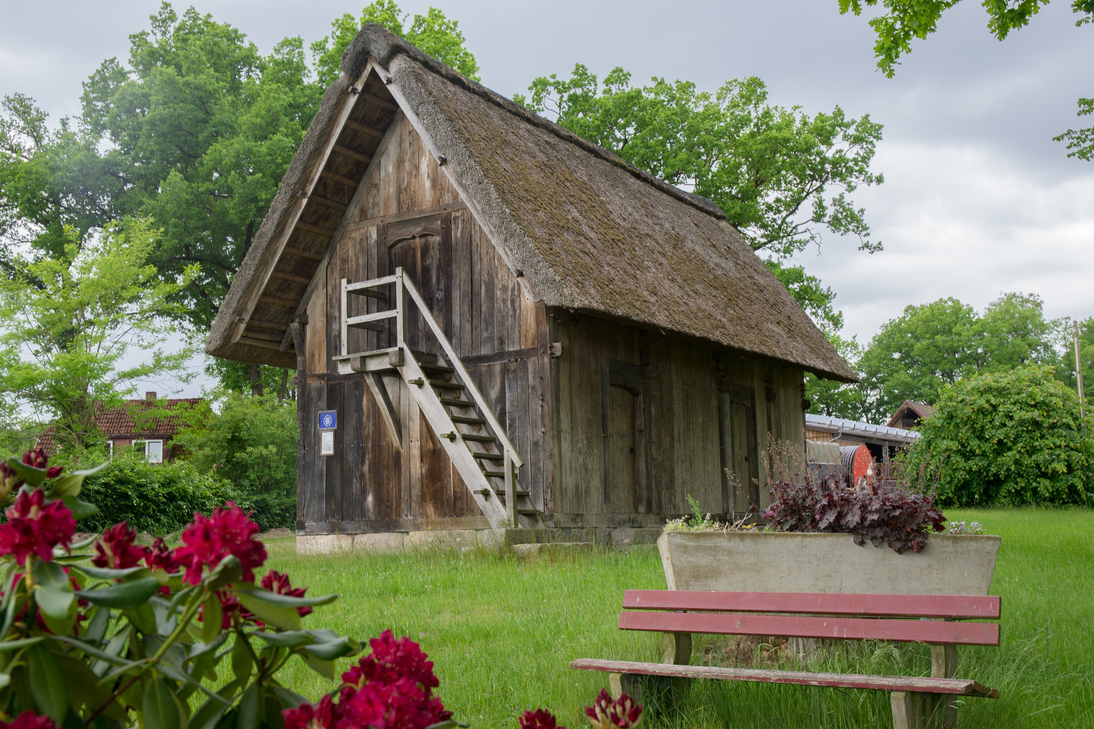 Historischer Treppenspeicher mit Reetdach auf grüner Wiese, umgeben von Bäumen und Blumen.
