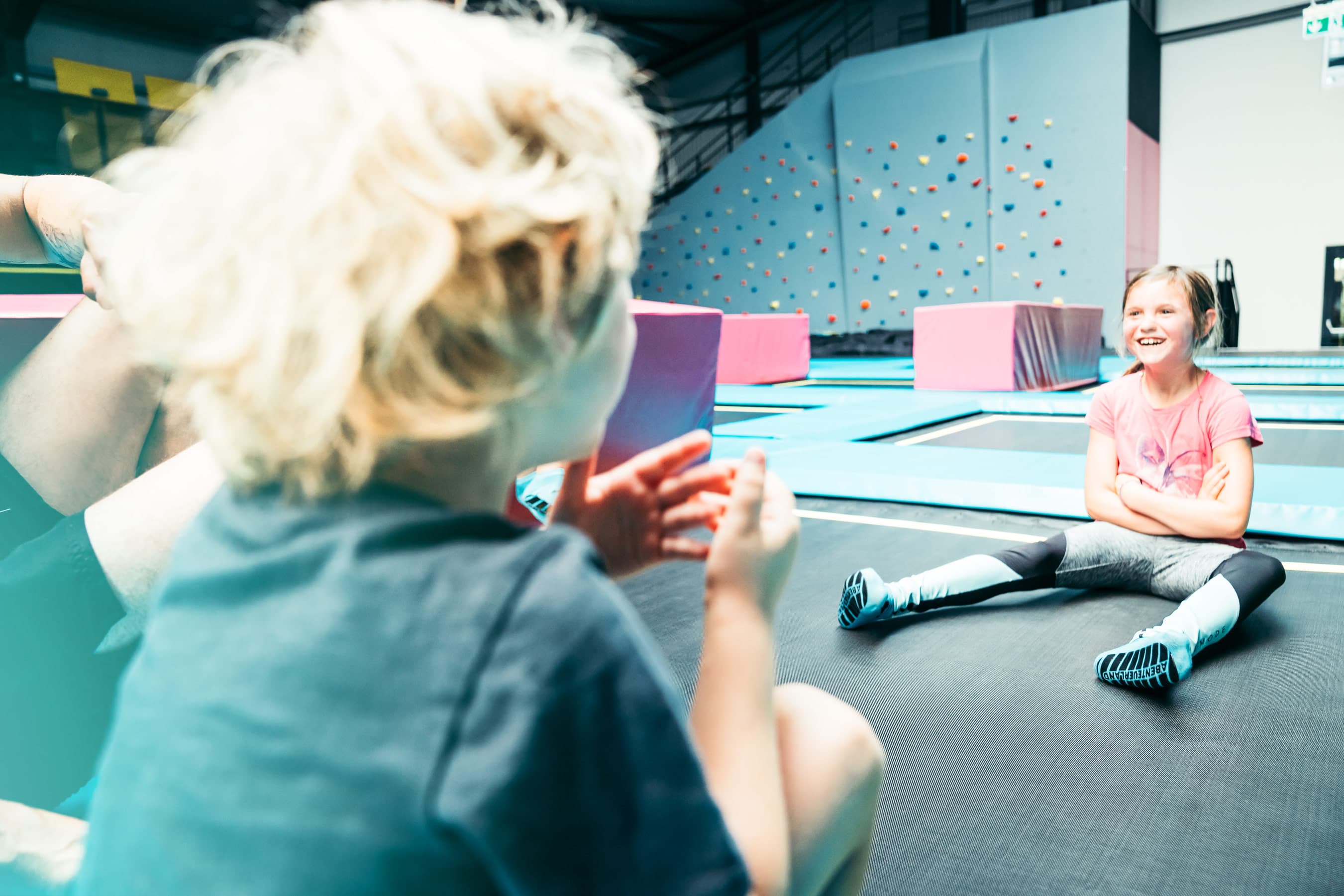 Ein blondes Kind im Fokus sitzt auf einem Trampolin, während ein lächelndes Mädchen im Hintergrund auf dem Trampolin entspannt. Im Raum sind bunte Plattformen und eine Kletterwand zu sehen.