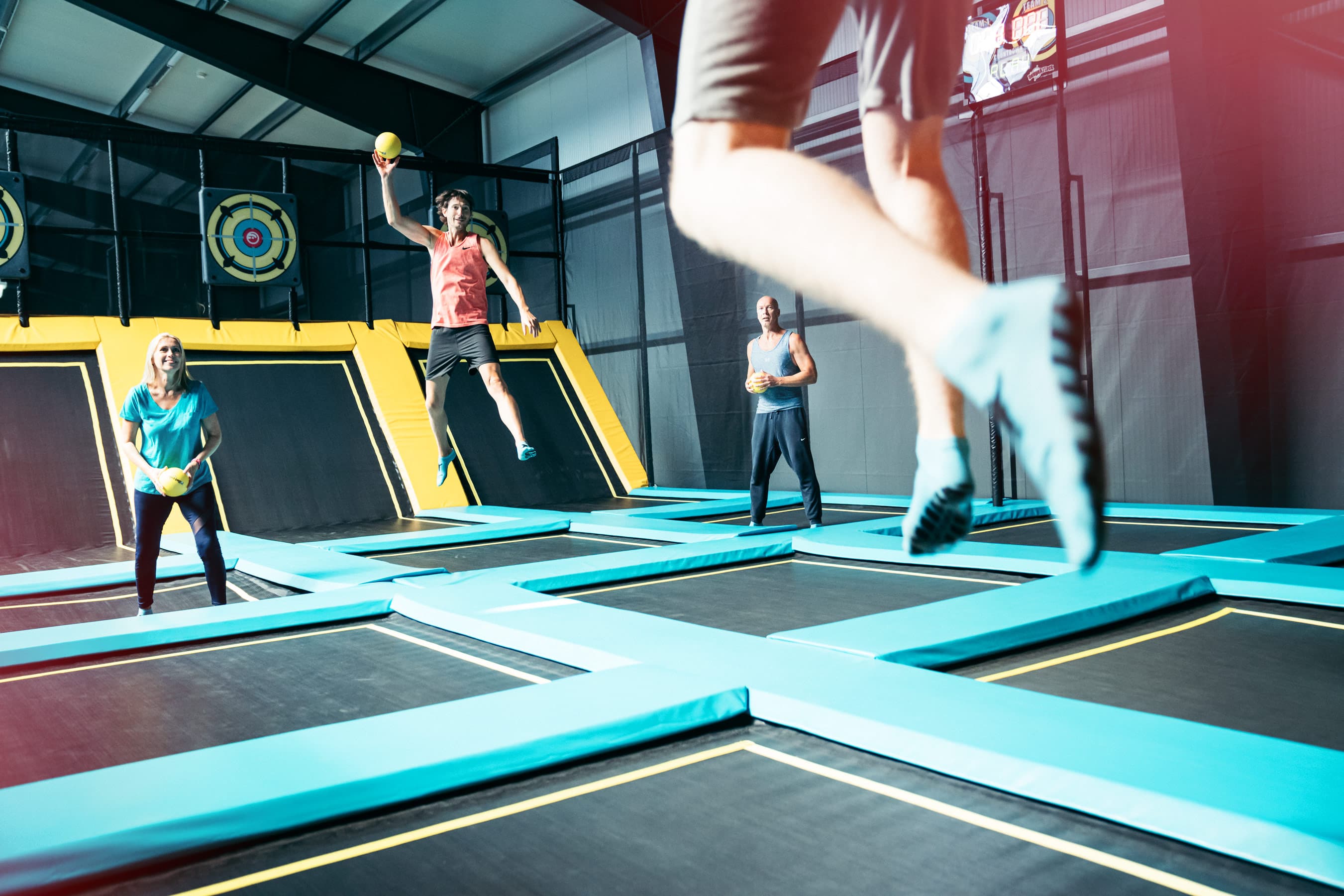 Eine Gruppe von Menschen hat Spaß auf einem Trampolinpark mit bunten Sprungflächen und Zielscheiben. Eine Person springt mit ausgestrecktem Arm und Ball in der Hand.