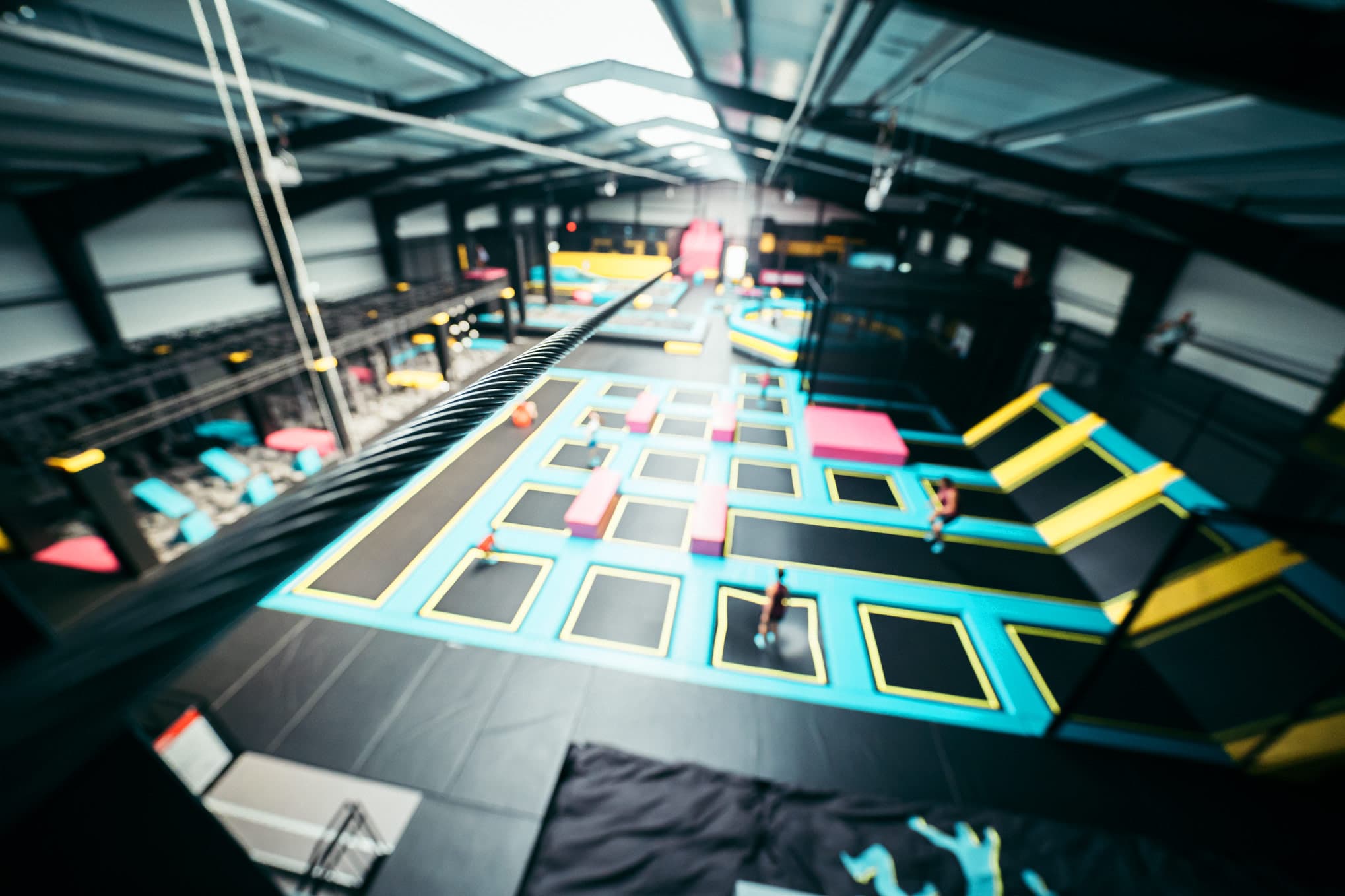 Das Bild zeigt eine große, helle Halle mit verschiedenen Trampolinen in leuchtenden Farben, die zur sportlichen Nutzung angeordnet sind. Bewegung im Hintergrund deutet auf aktive Besucher hin.