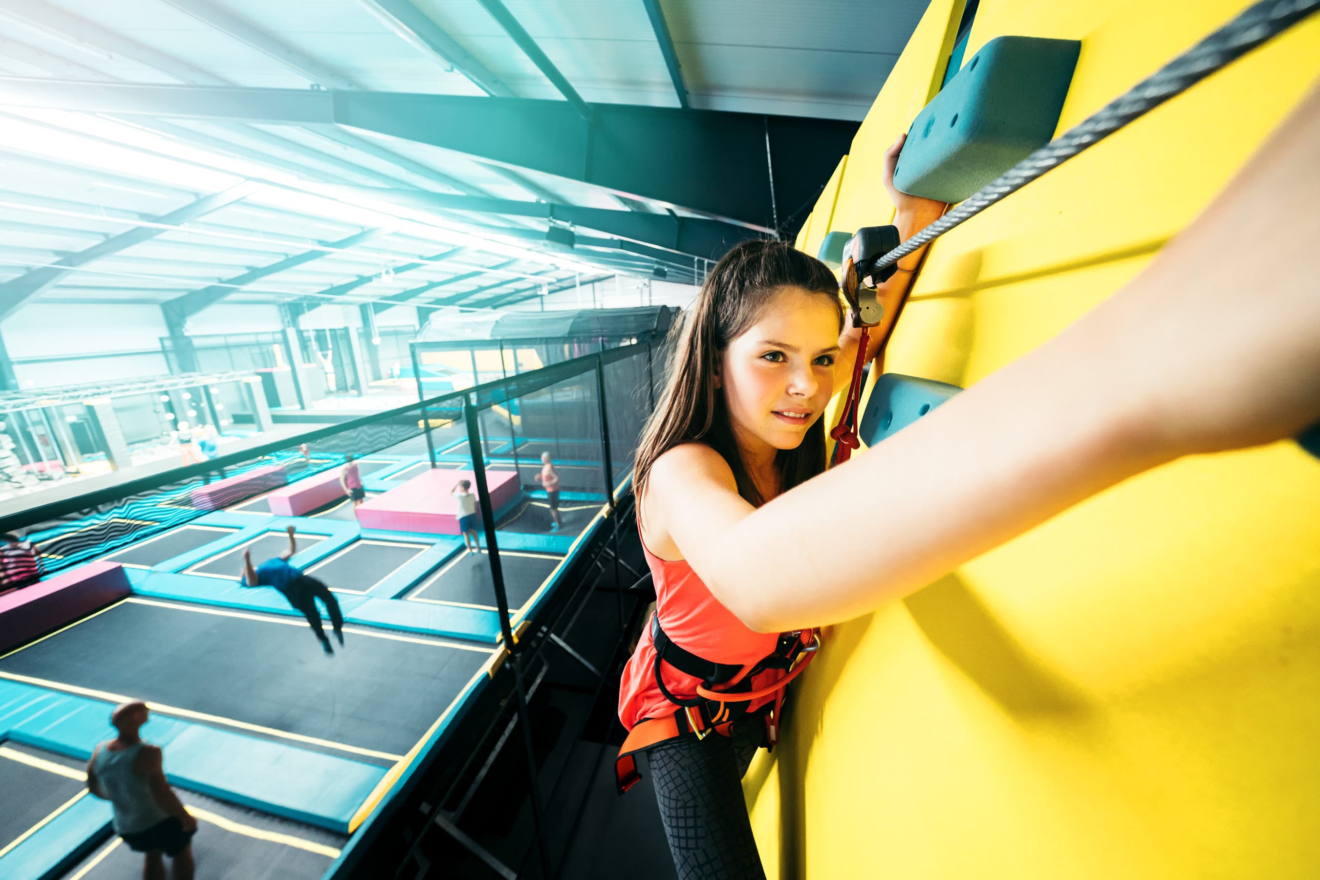 Eine junge Frau erklimmt konzentriert eine gelbe Kletterwand, umgeben von Trampolin-Arealen.