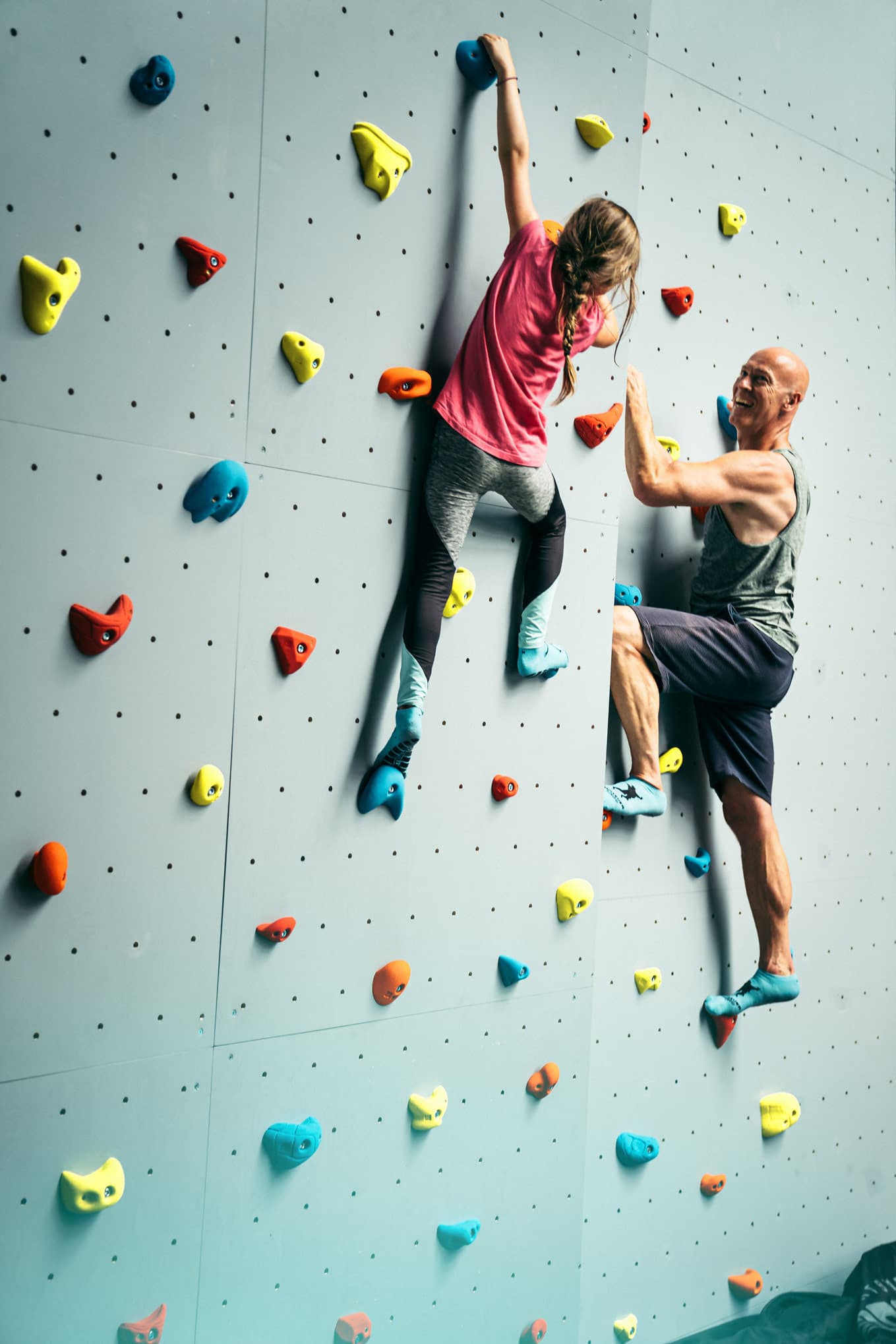 Ein Mädchen und ein Mann klettern an einer bunten Kletterwand, fröhlich und konzentriert.