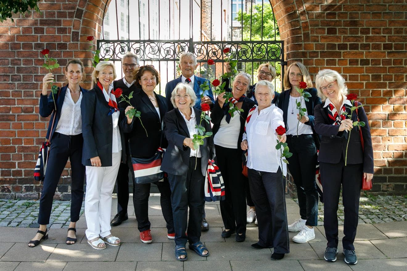 Zehn lachende Rote Rosen Stadtführer:innen halten rote Rosen vor einem Backsteintor.