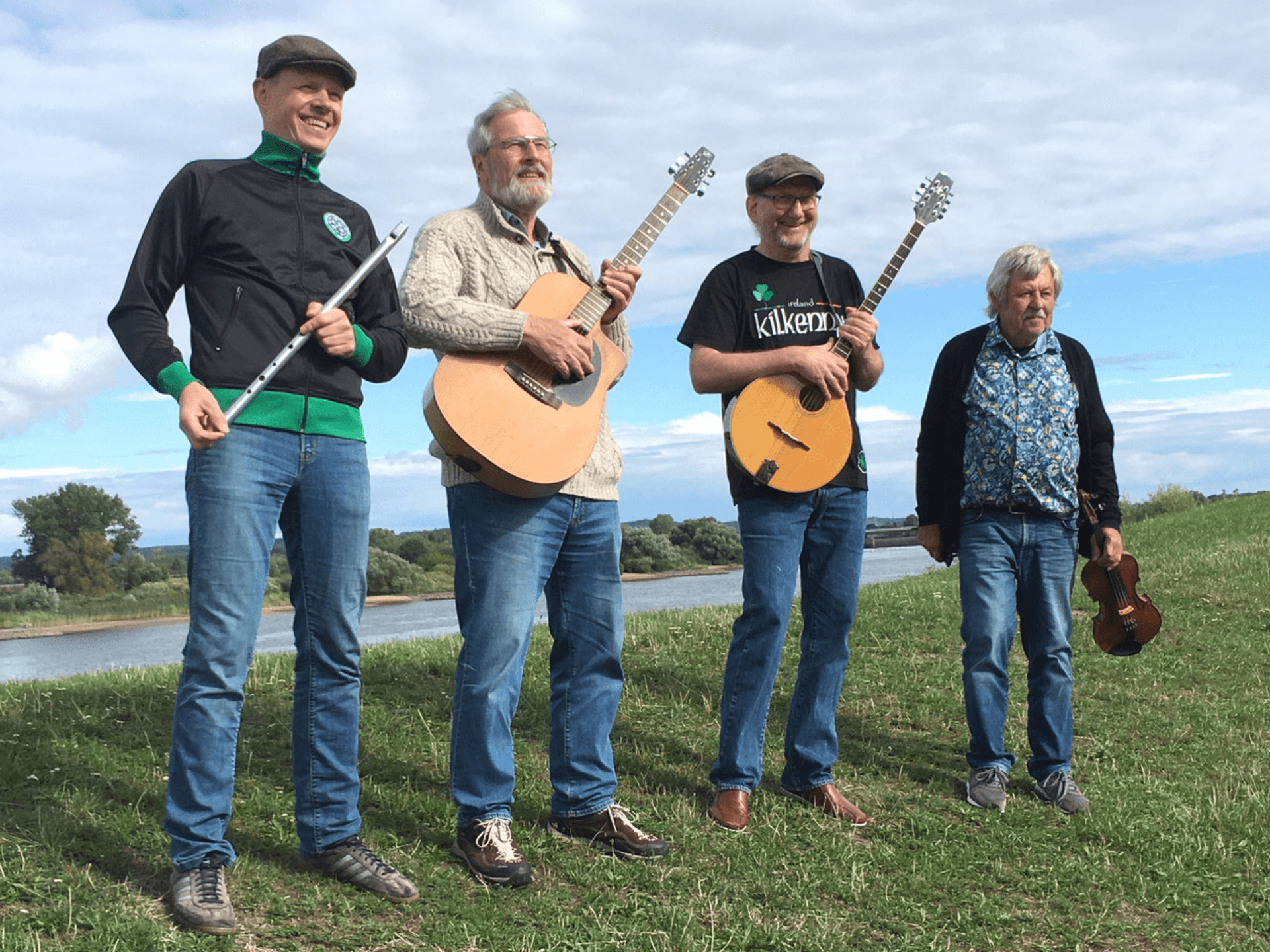 Vier Musiker mit Instrumenten stehen im Freien vor einer Flusslandschaft und lachen in die Kamera.