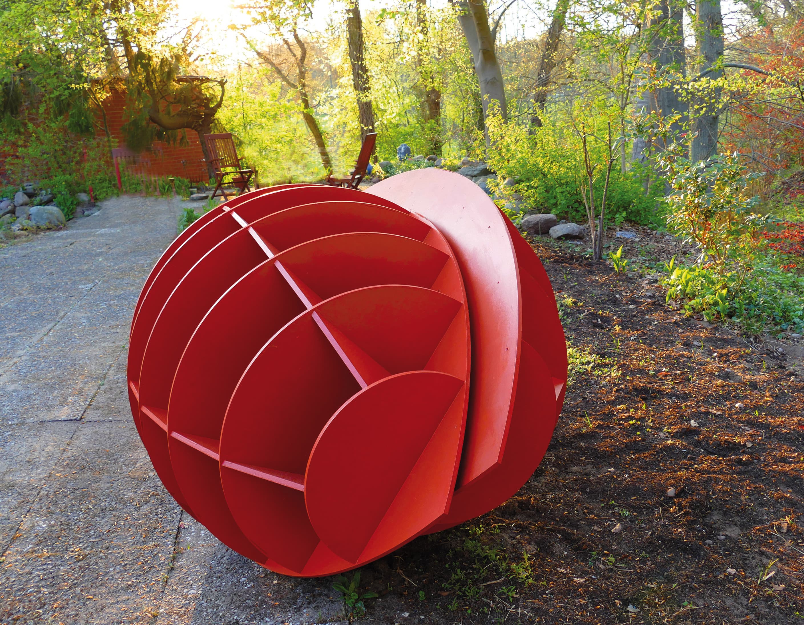 Das Bild zeigt eine kunstvolle, rot bemalte Skulptur im Freien, die kugelförmig gestaltet ist und aus mehreren Segmenten besteht. Sie liegt auf einem gepflasterten Weg und wird von grüner Vegetation umrahmt.