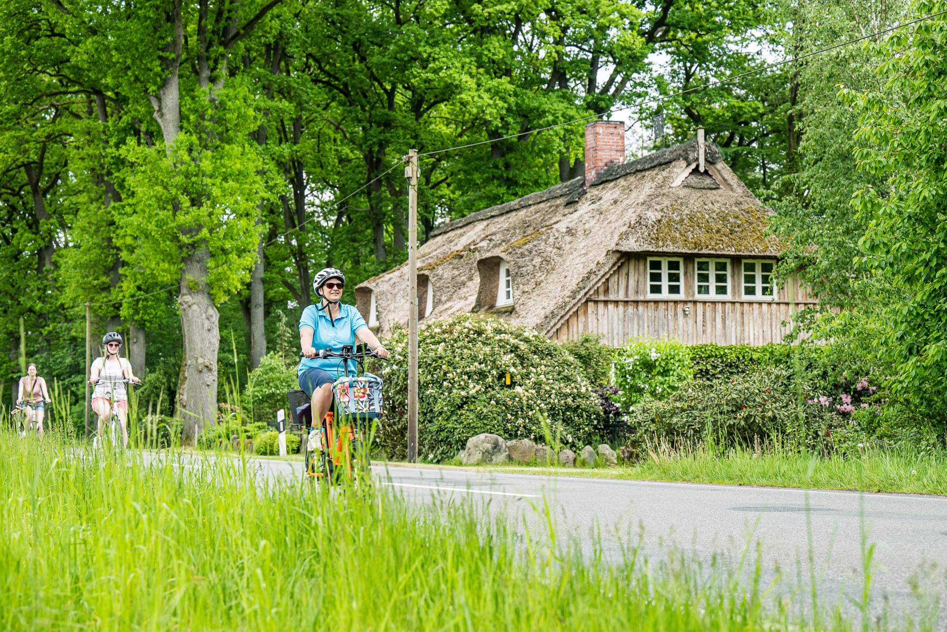 Drei Frauen radeln an einem reetgedeckten Bauernhaus unter grünen Bäumen vorbei.