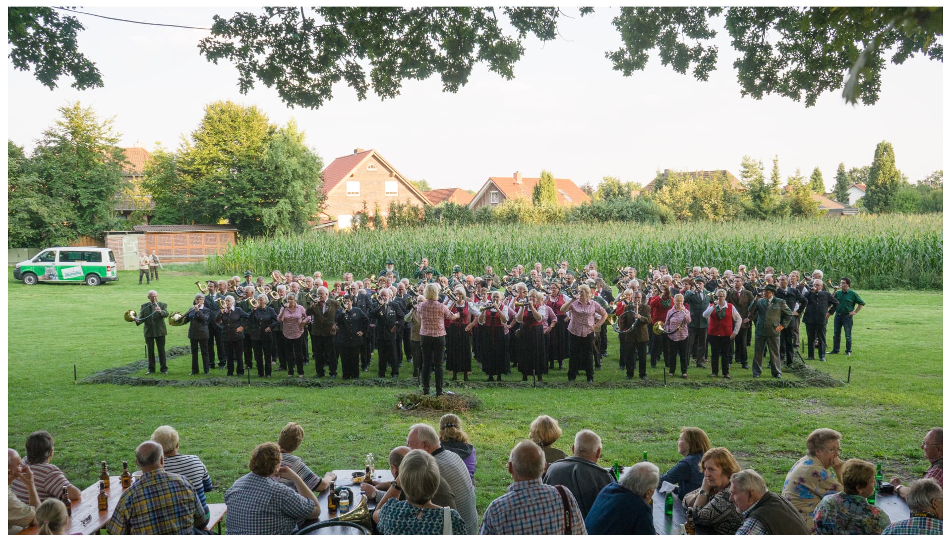 Eine große Gruppe in Trachten spielt im Freien Blasmusik vor einem Publikum bei einem Fest.