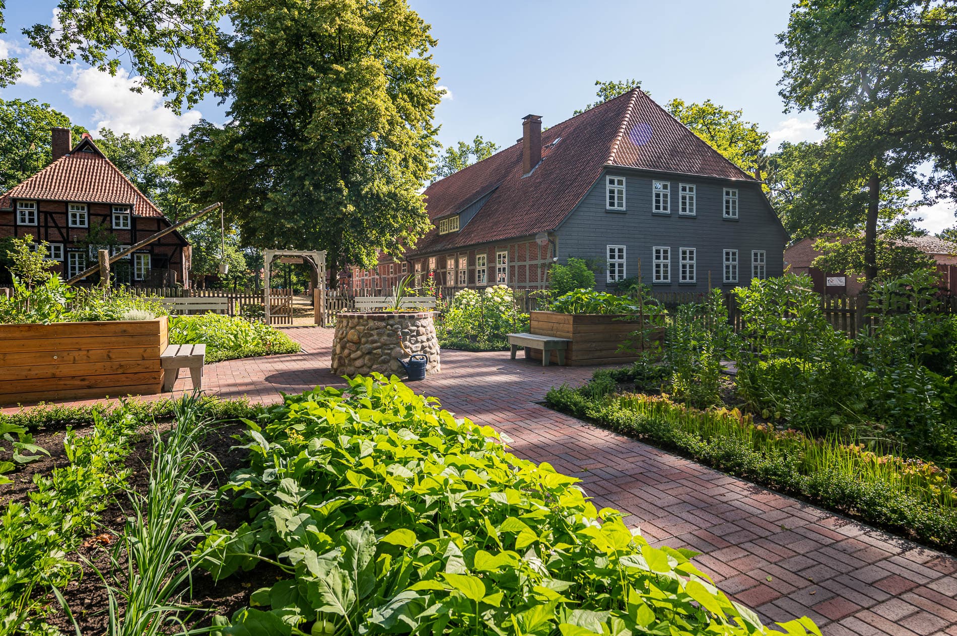 Ein gepflegter Kräutergarten mit Hochbeeten aus Holz. Im Hintergrund steht ein traditionelles Bauernhaus mit rotem Ziegeldach und Fachwerkfassade. Der Garten ist umgeben von hohen Bäumen, die Schatten spenden. Ein gepflasterter Weg schlängelt sich durch das Grün, und die Pflanzen wirken gesund und kräftig.
