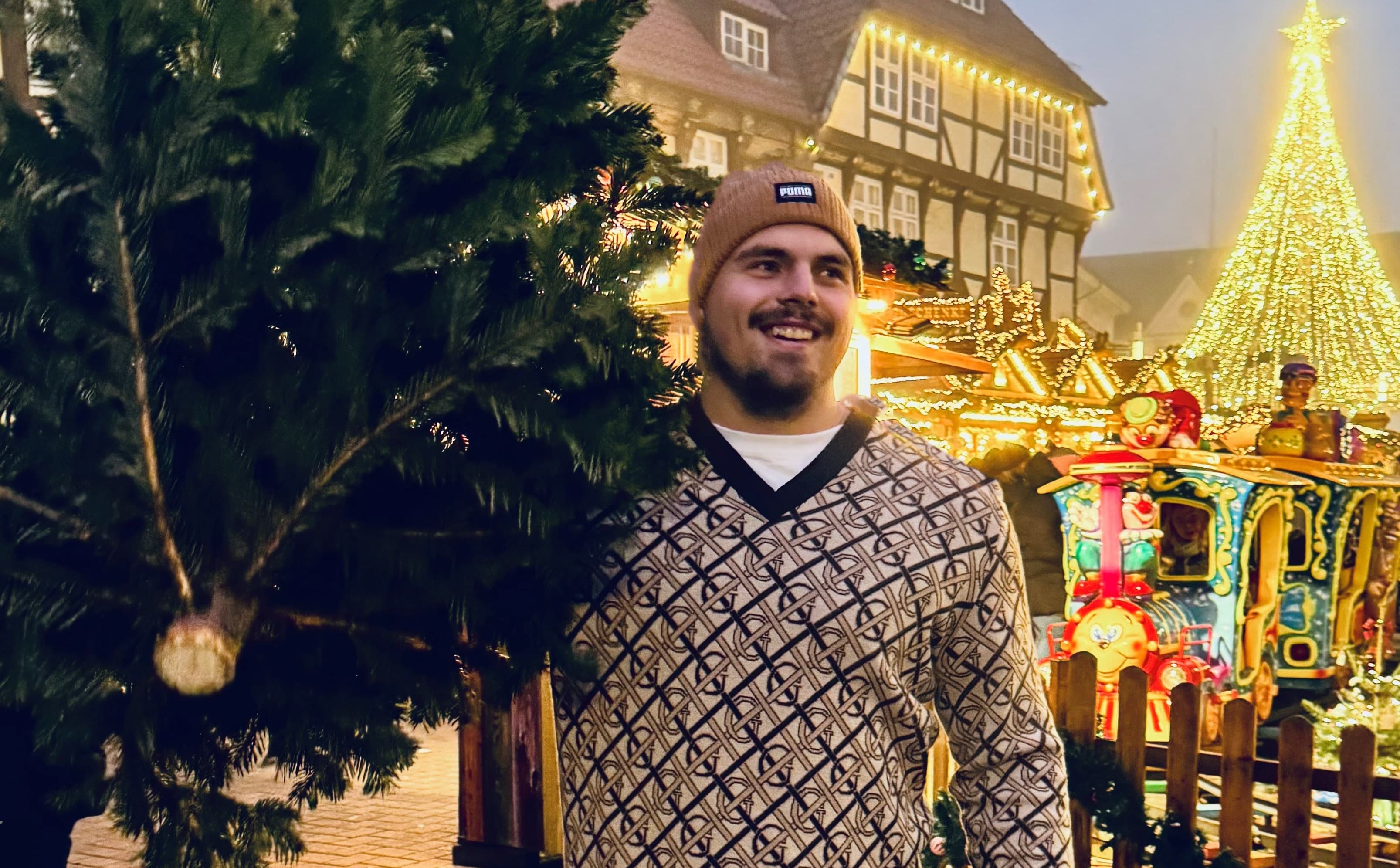 Max Dehning mit festlichem Hintergrund auf Weihnachtsmarkt, lächelnd mit Tanne.Max Dehning with festive background at Christmas market, smiling with fir tree.Max Dehning med festlig baggrund på julemarked, smilende med grantræ.Max Dehning met feestelijke achtergrond op kerstmarkt, lachend met dennenboom.