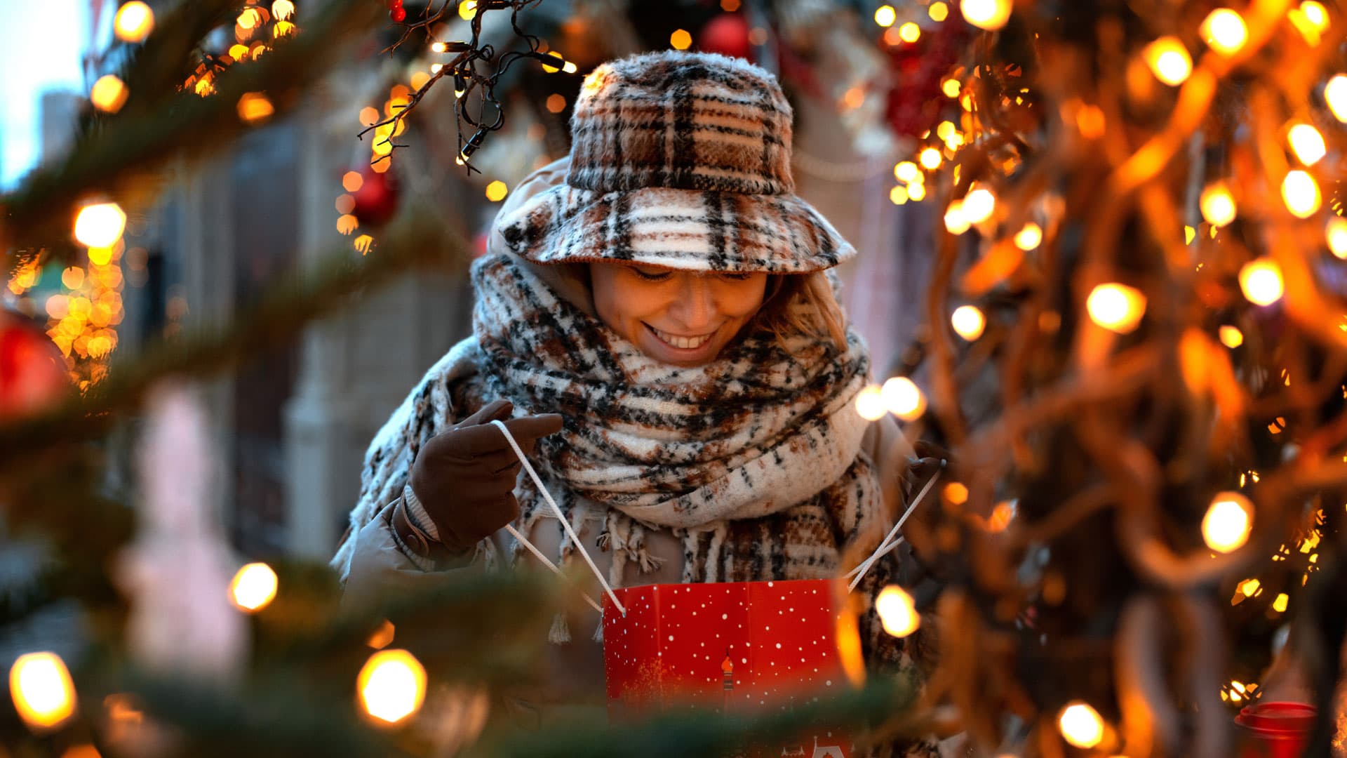 Eine junge Frau, warm eingepackt mit Schal und Hut, öffnet gespannt eine rote Geschenktüte.A young woman, wrapped up warmly in a scarf and hat, eagerly opens a red gift bag.En ung kvinde, varmt pakket ind i halstørklæde og hue, åbner ivrigt en rød gavepose.Een jonge vrouw, warm ingepakt in een sjaal en muts, opent gretig een rode geschenktas.