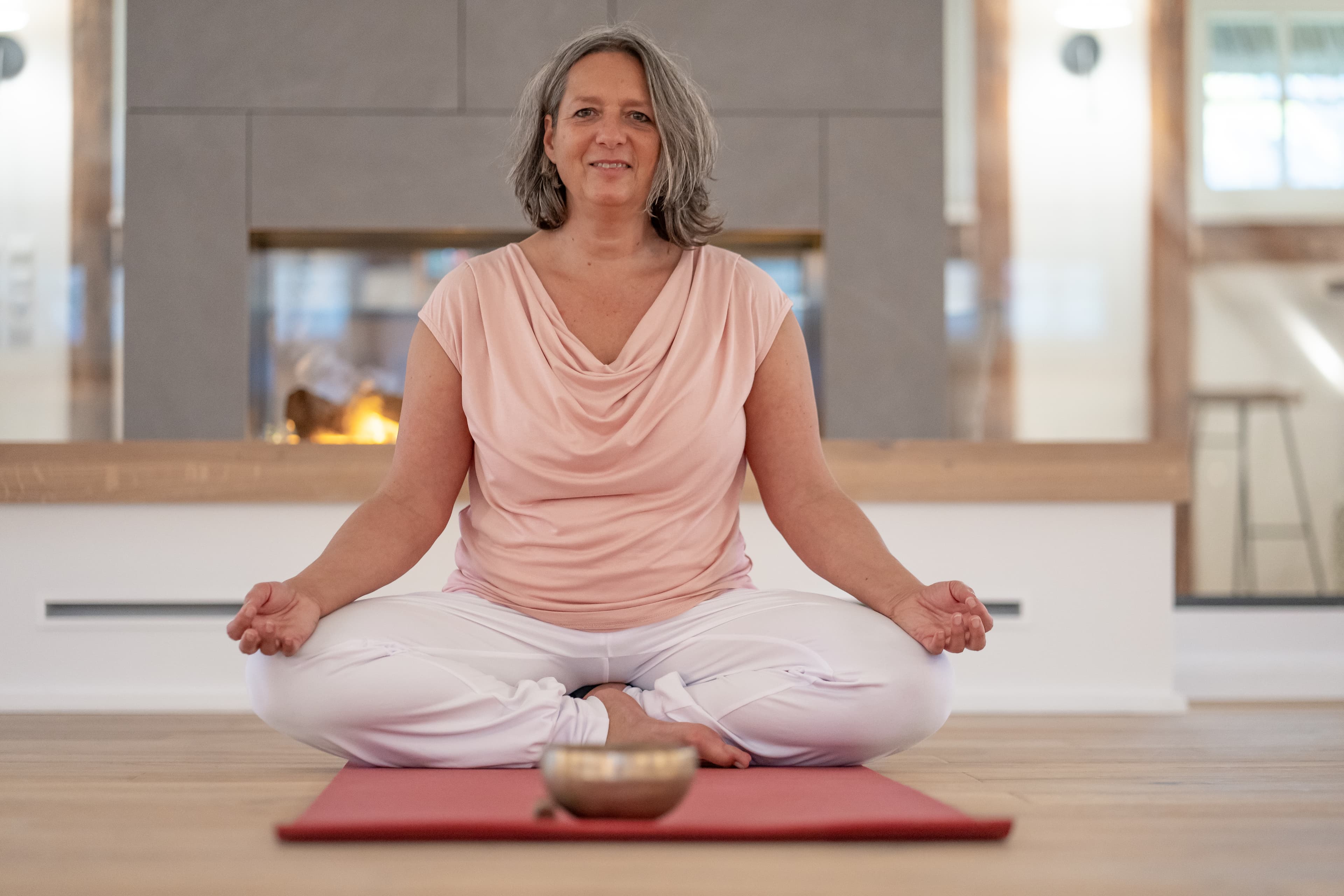 Frau sitzt entspannt im Schneidersitz auf einer Yogamatte vor einem Kamin und meditiert.