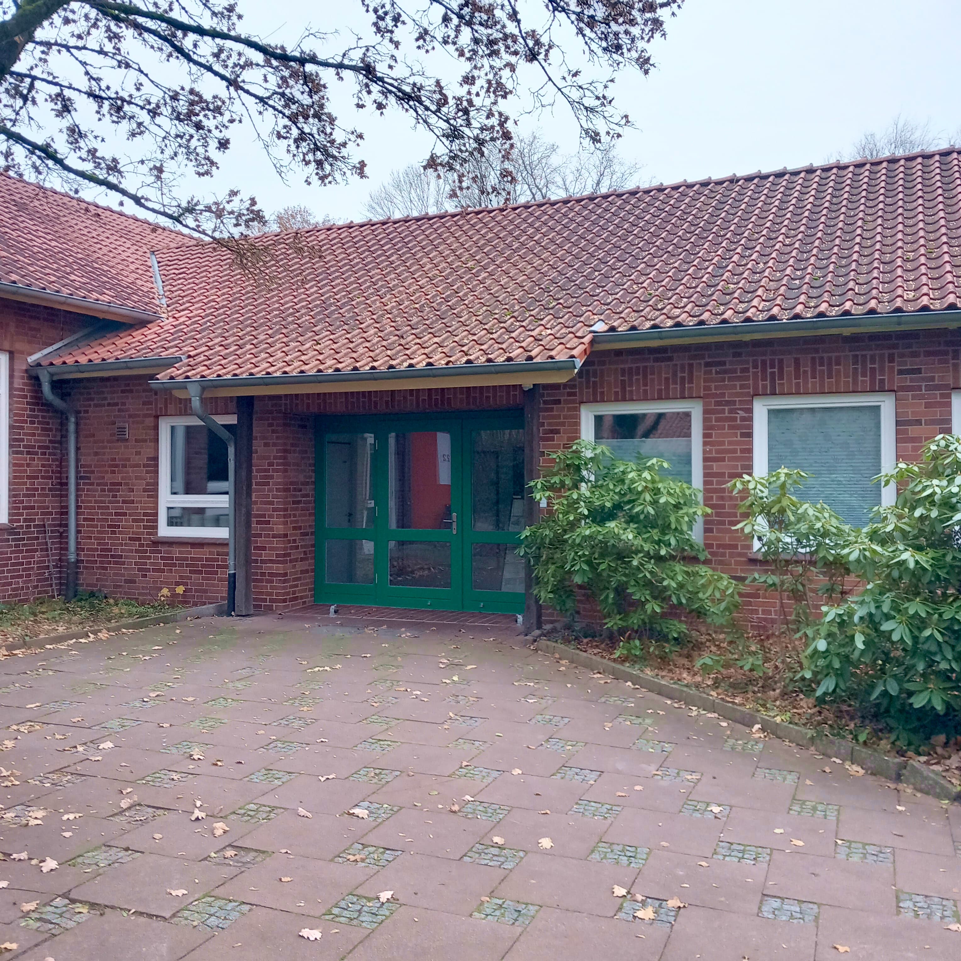 Backsteingebäude mit rotem Ziegeldach, begrüntem Eingang und gepflastertem Vorplatz.