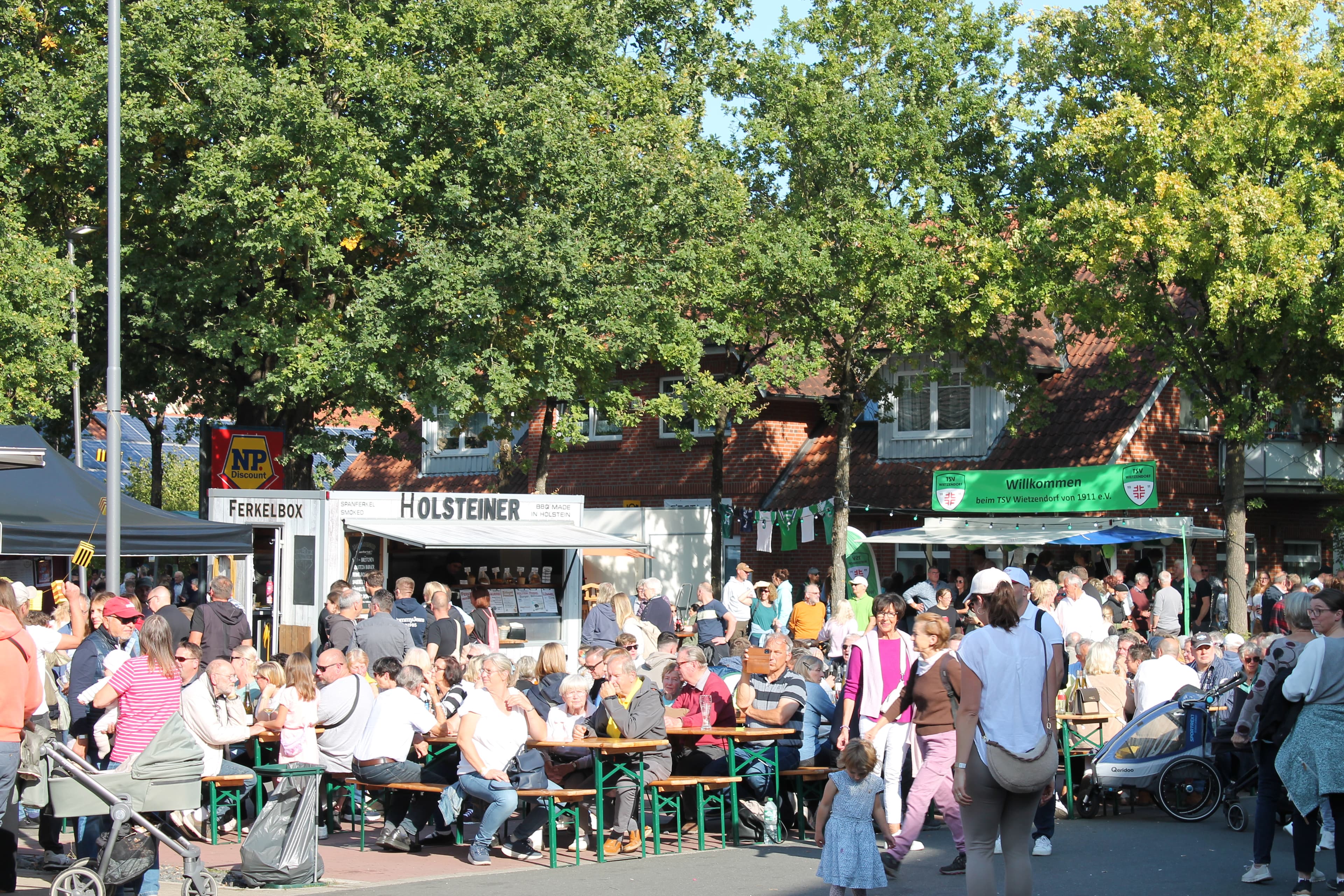 Menschen sitzen und stehen bei sonnigem Wetter auf dem Festplatz vor der Bühne beim Honigfest in Wietzendorf.People sit and stand in sunny weather on the fairground in front of the stage at the honey festival in Wietzendorf.Folk sidder og står i solskinsvejr på markedspladsen foran scenen på honningfestivalen i Wietzendorf.Mensen zitten en staan bij zonnig weer op de kermis voor het podium op het honingfestival in Wietzendorf.