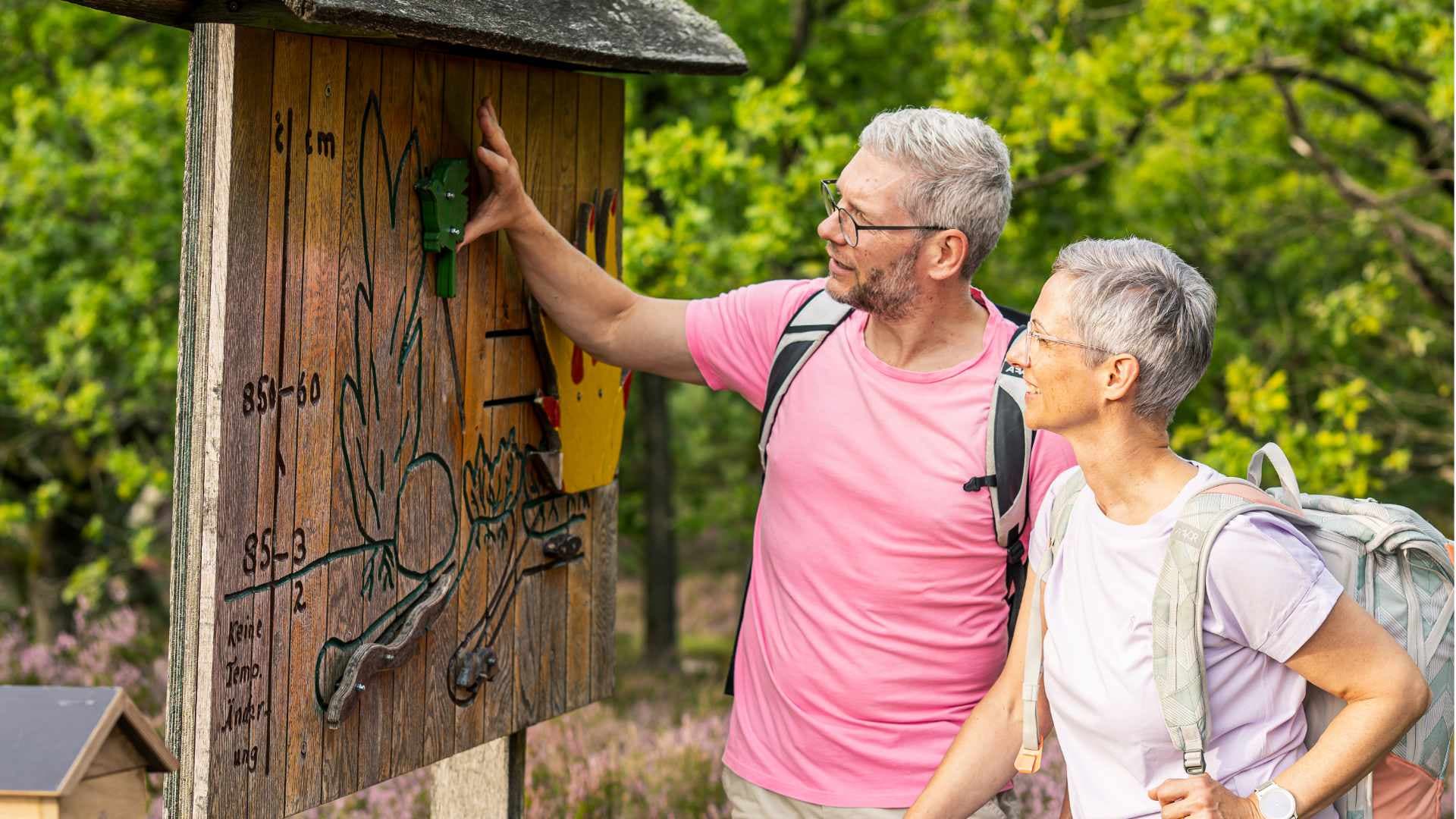 Wanderndes Paar betrachtet interaktive Holztafel am Machandel-Erlebnispfad im Naturschutzgebiet.
