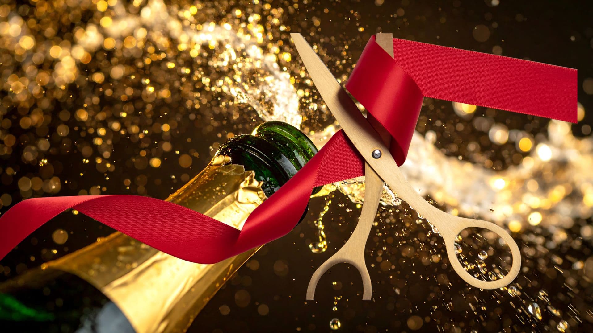 Goldene Schere durchschneidet ein rotes Band vor einer sprudelnden Sektflasche.