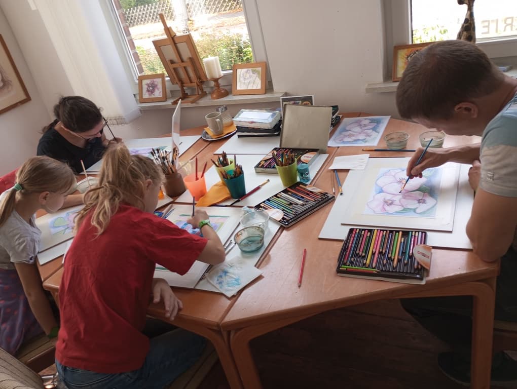 Erwachsene und Kinder malen konzentriert mit Stiften und Farben an einem Tisch im Atelier.