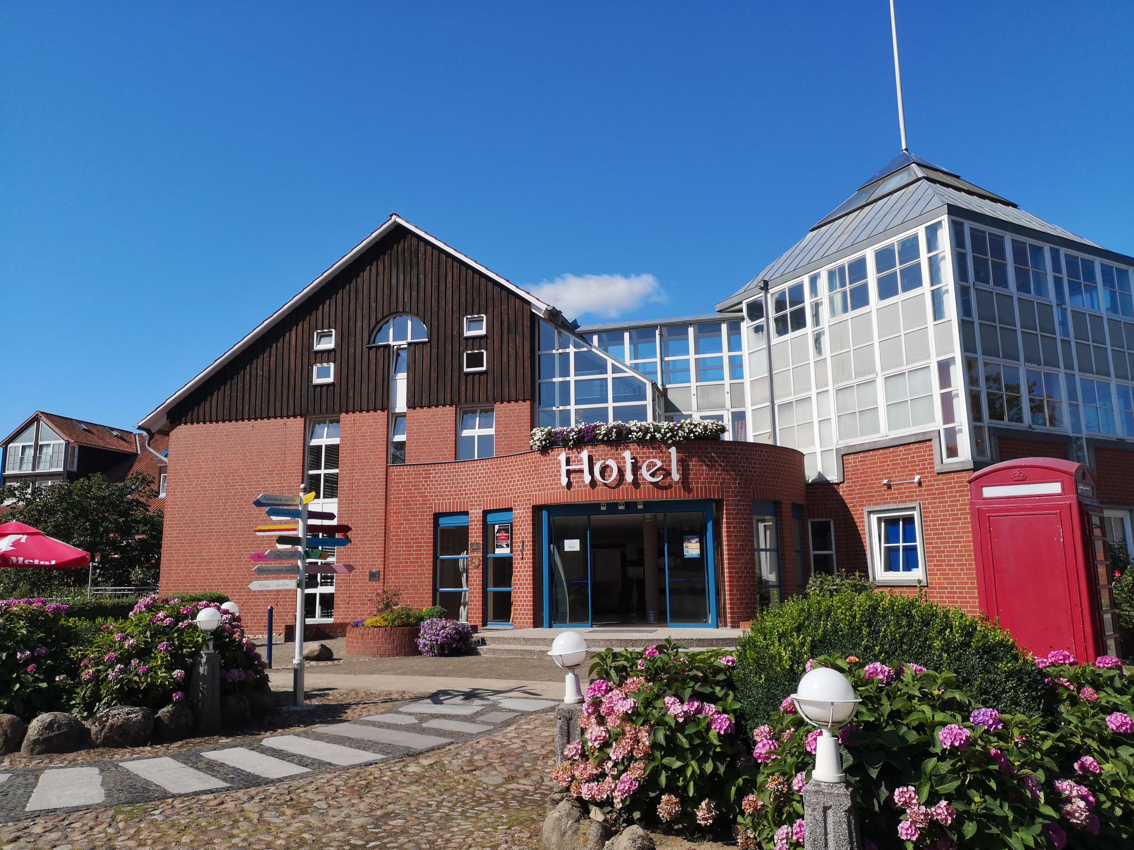 Außenansicht des Heide Hotel Reinstorf mit roter Backsteinfassade, blauem Himmel und blühenden Hortensien.