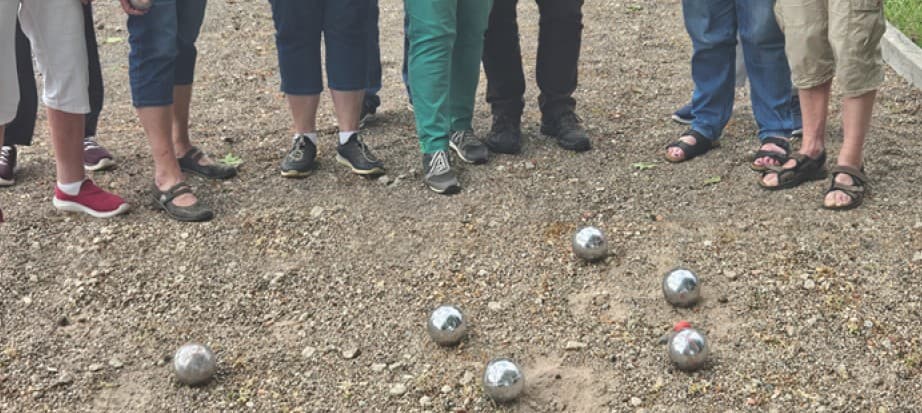 Eine Grupperuppe von Menschen, nur die Beine und Füße sind zu sehen, steht im Halbkreis um glänzende Boulekugeln auf einem Schotterplatz.