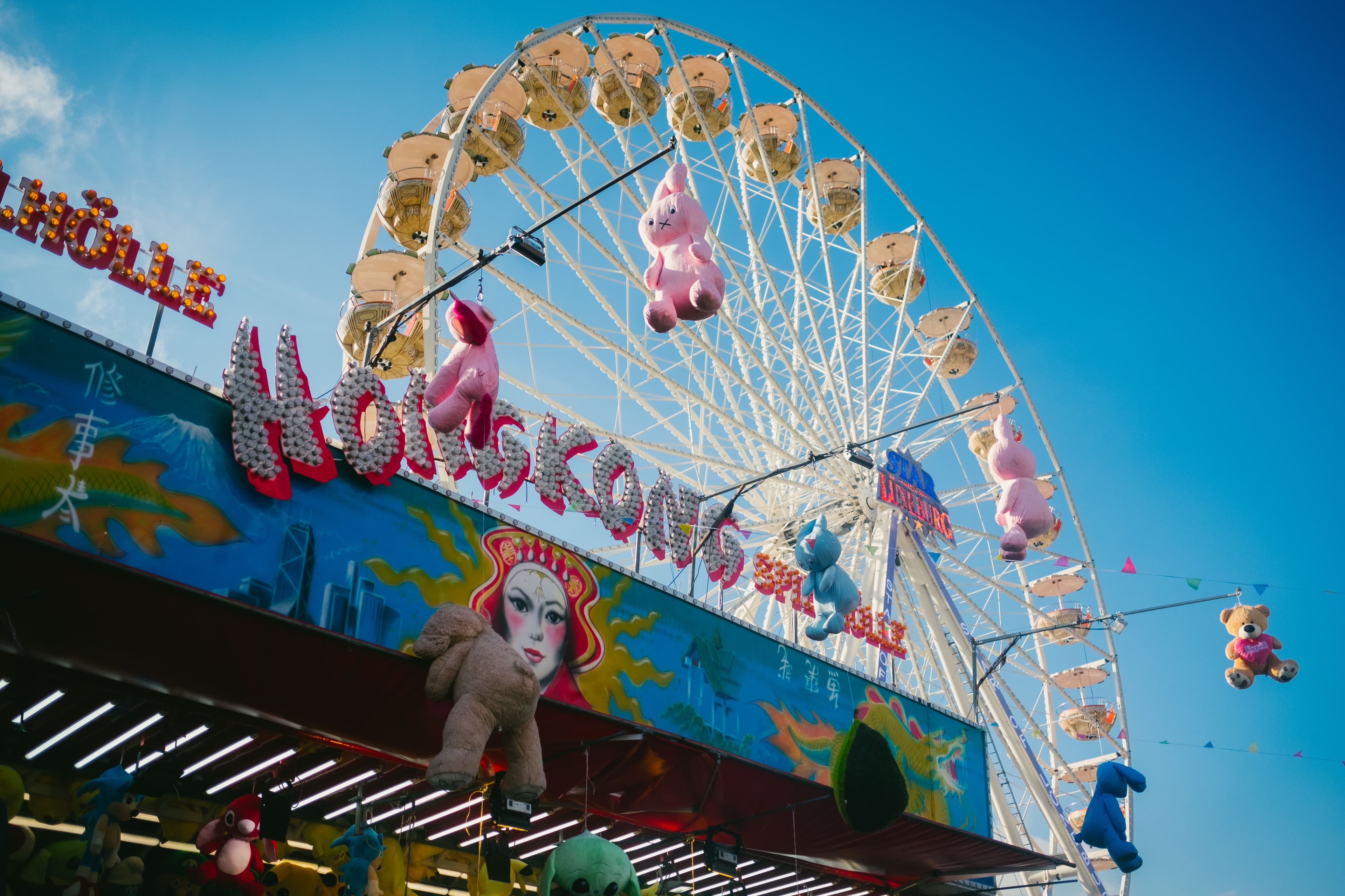 Bunte Jahrmarktsbude mit Plüschtieren vor einem großen Riesenrad unter blauem Himmel auf dem Lüneburger Frühjahrsmarkt.