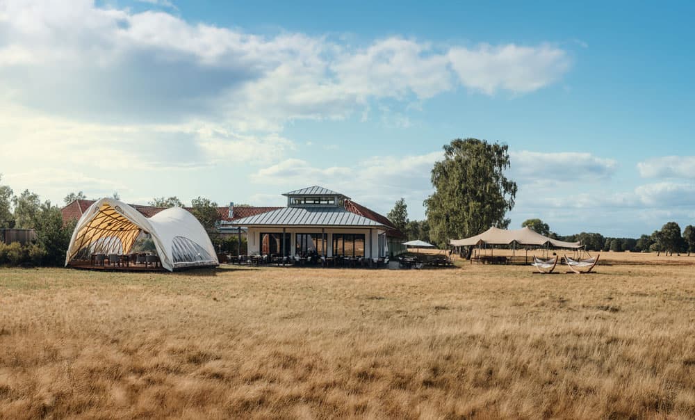 Strohboid-Pavillon und Gebäude auf einer weiten trockenen Grasfläche unter blauem Himmel.