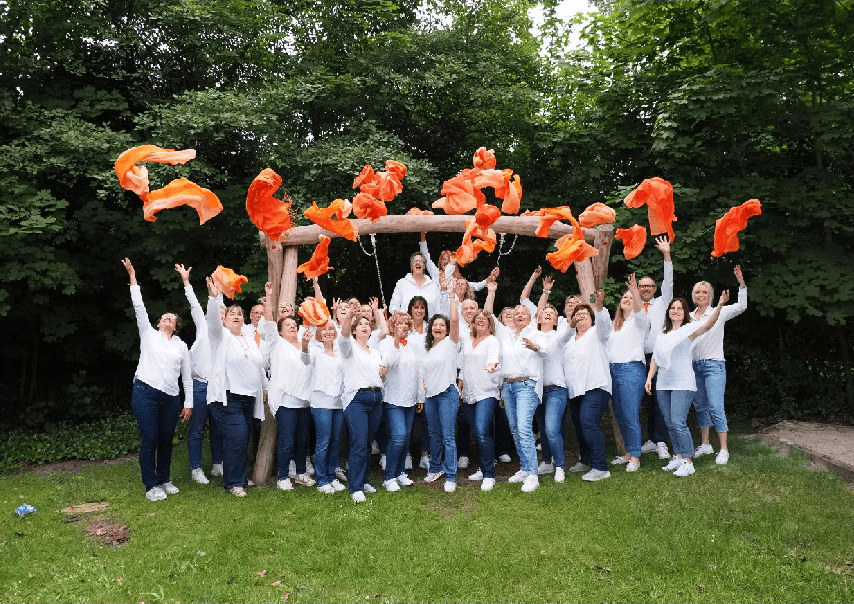 Ein Chor in weißen Hemden und Jeans wirft im Grünen jubelnd orange Tücher hoch.