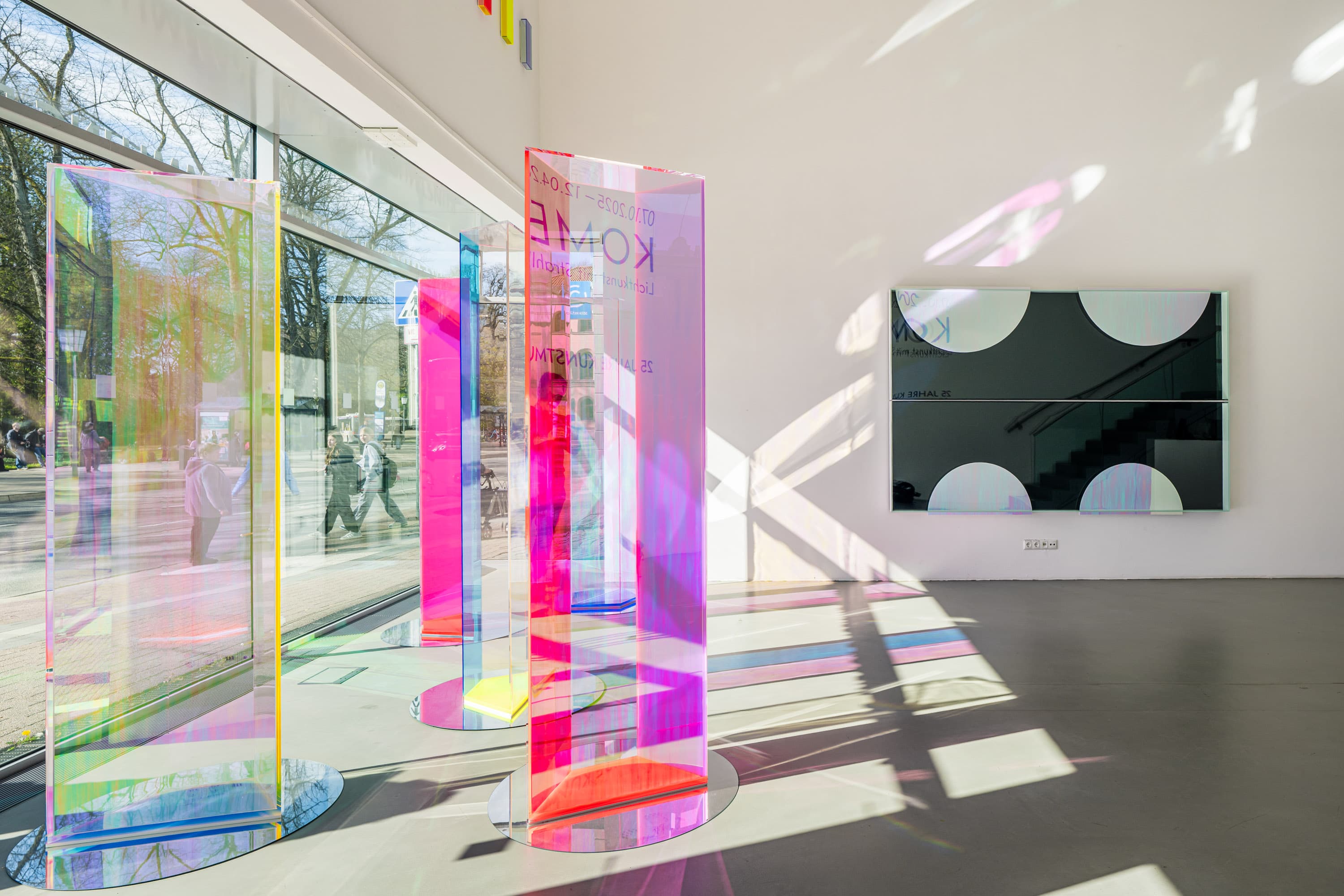 Lichtdurchfluteter Raum des Eingangsbereichs mit bunten, transparenten Stelen, die farbige Schatten werfen. Kunst von Regine Schumann