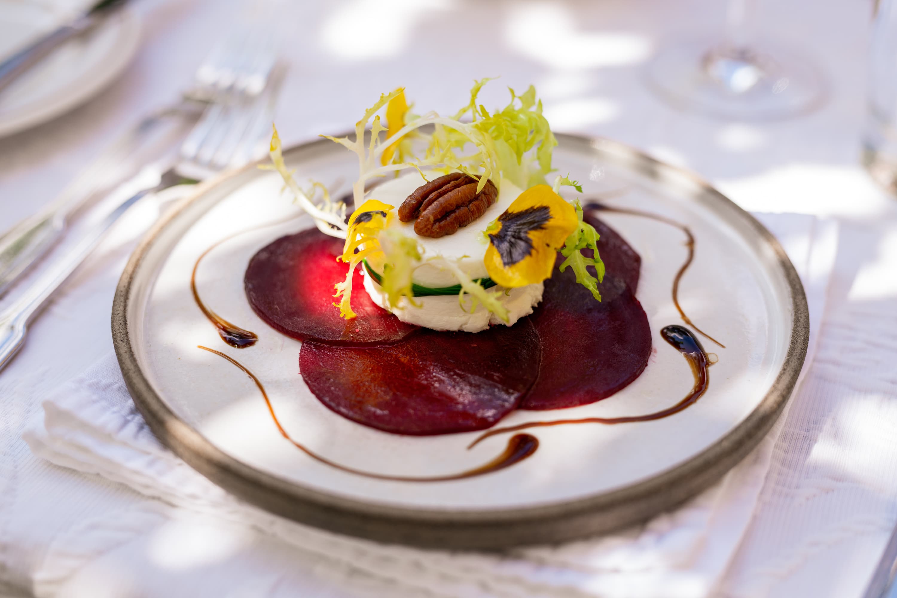 Rote-Beete-Carpaccio mit Ziegenkäse, Salat, Pekannuss und Blüte auf einem hellen Keramikteller.
