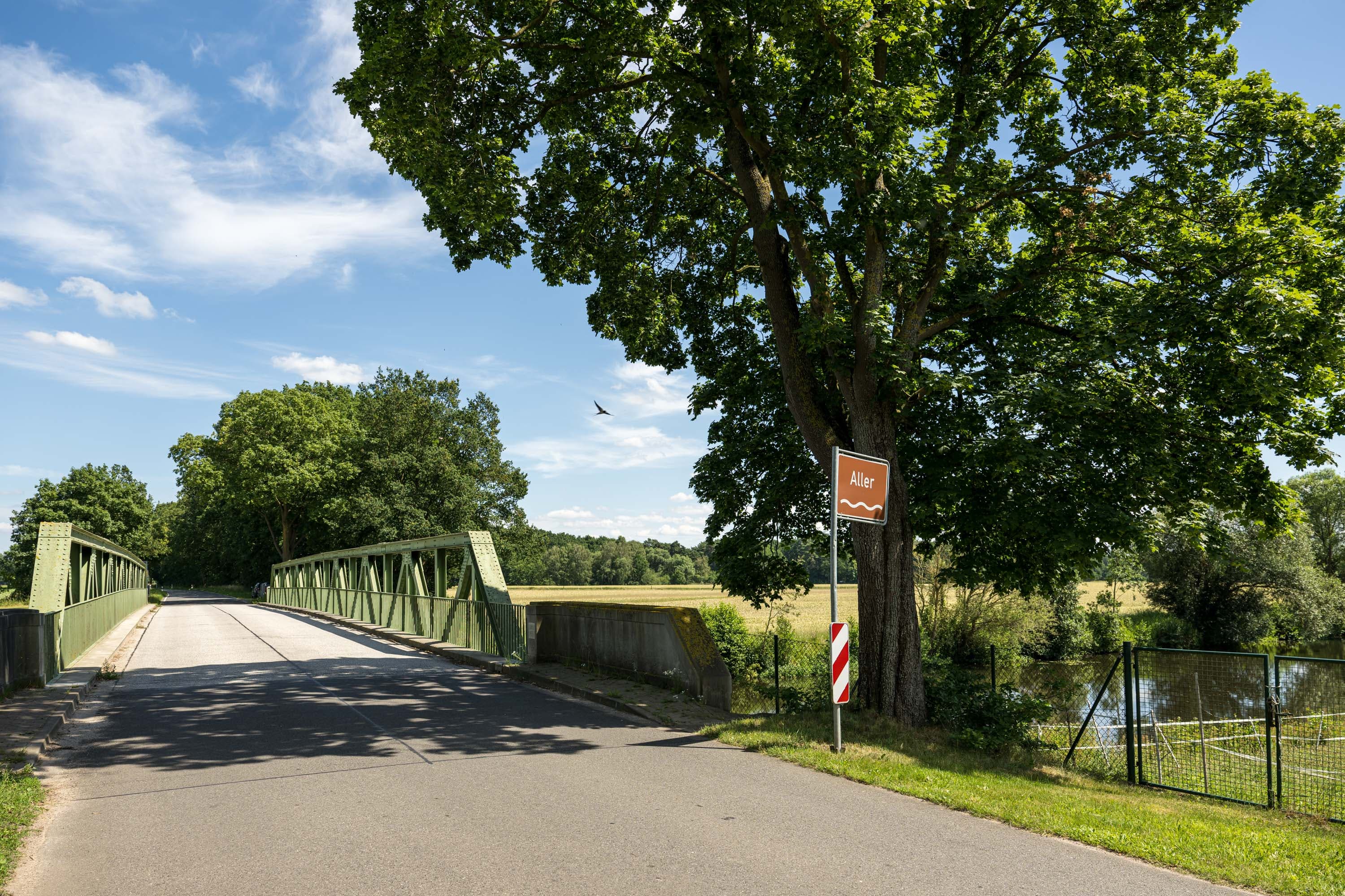 Grüne Brücke über die Aller bei Schwachhausen, daneben ein Flussschild unter großem Baum