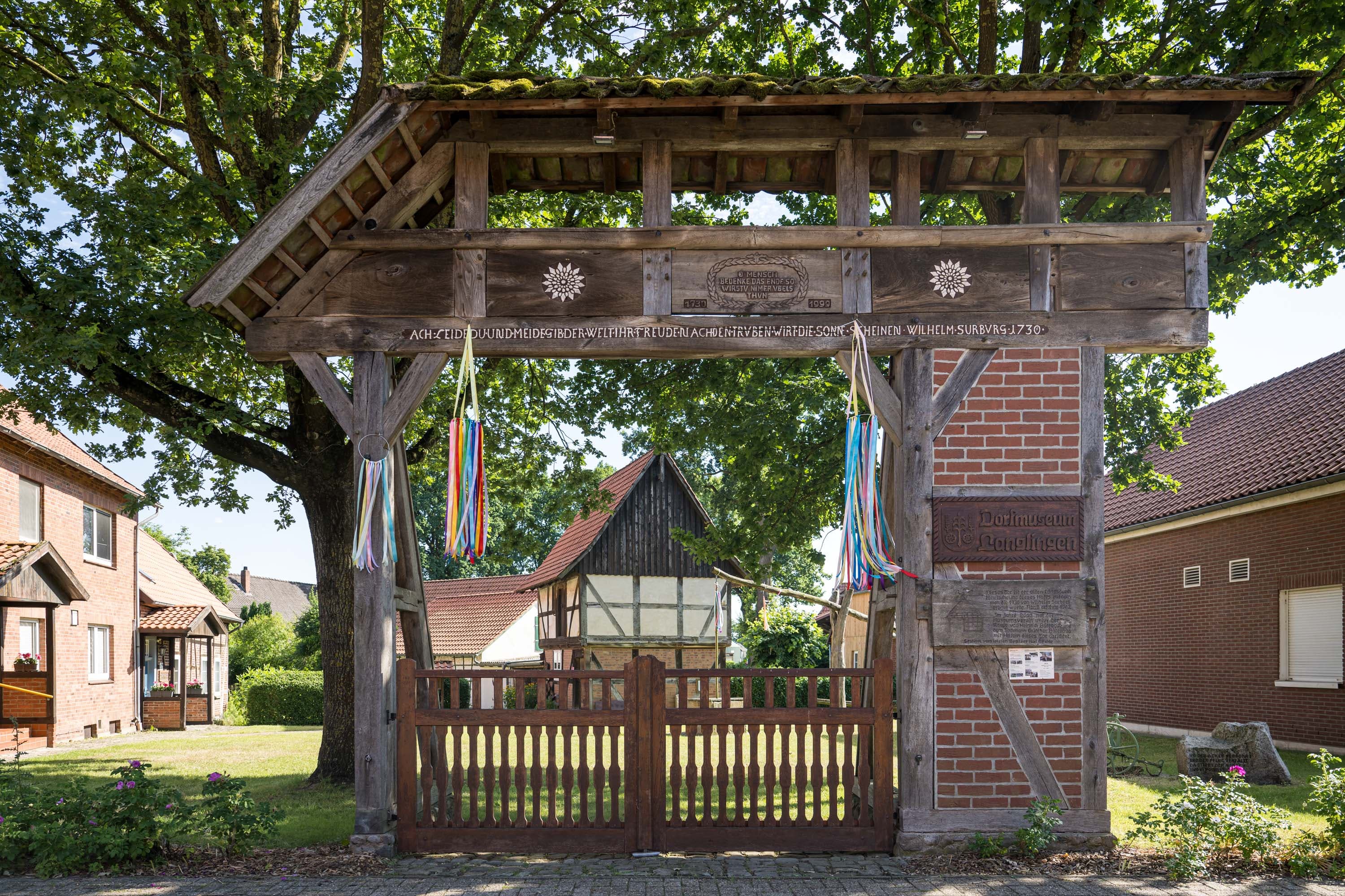 Holztor mit bunten Bändern am Dorfmuseum Langlingen, dahinter Fachwerkhaus und Bäume