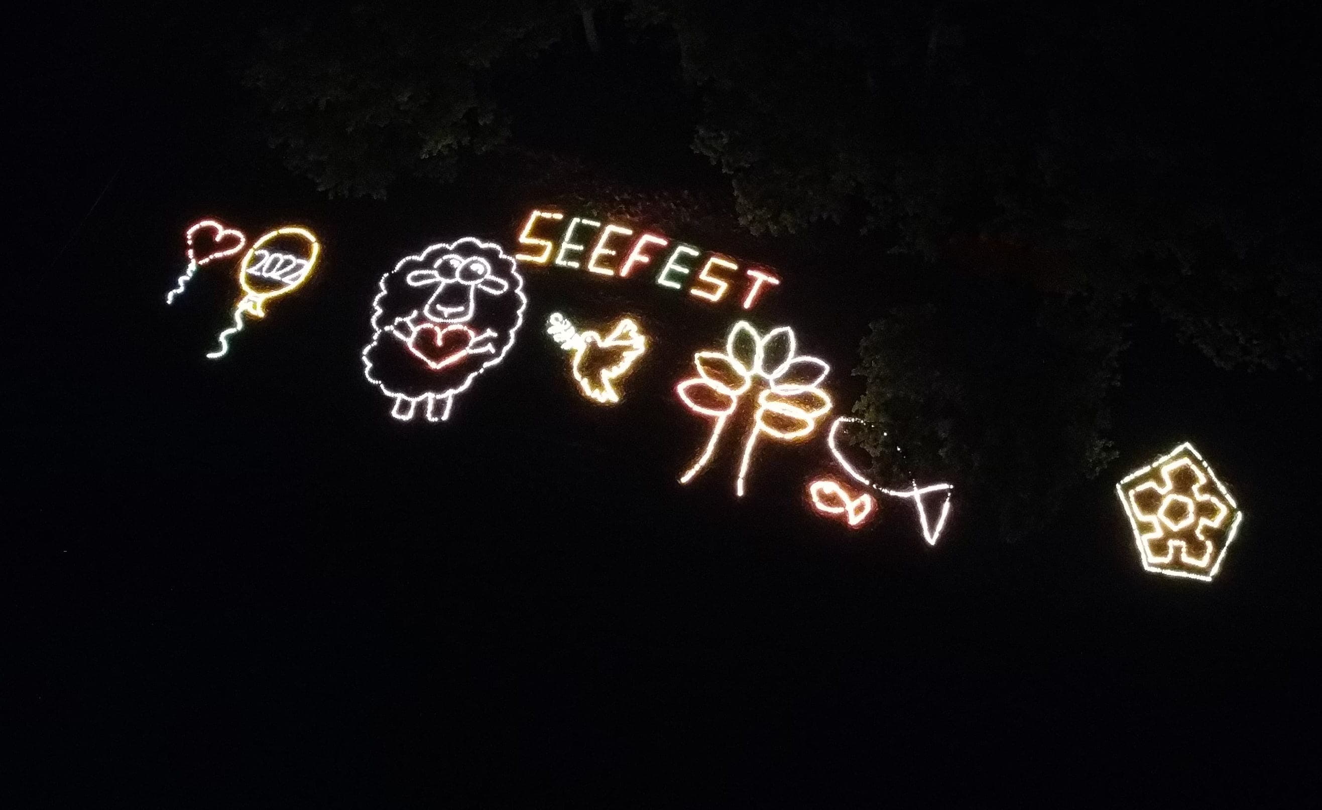 Leuchtfiguren mit Schriftzug Seefest strahlen nachts zwischen Baumkronen.