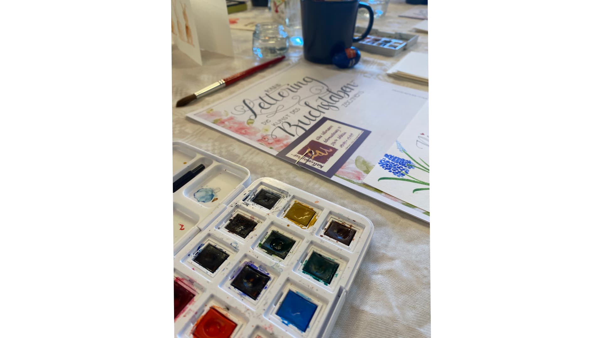 Aquarellfarben, Pinsel und Handlettering-Vorlage auf einem Workshop-Tisch.