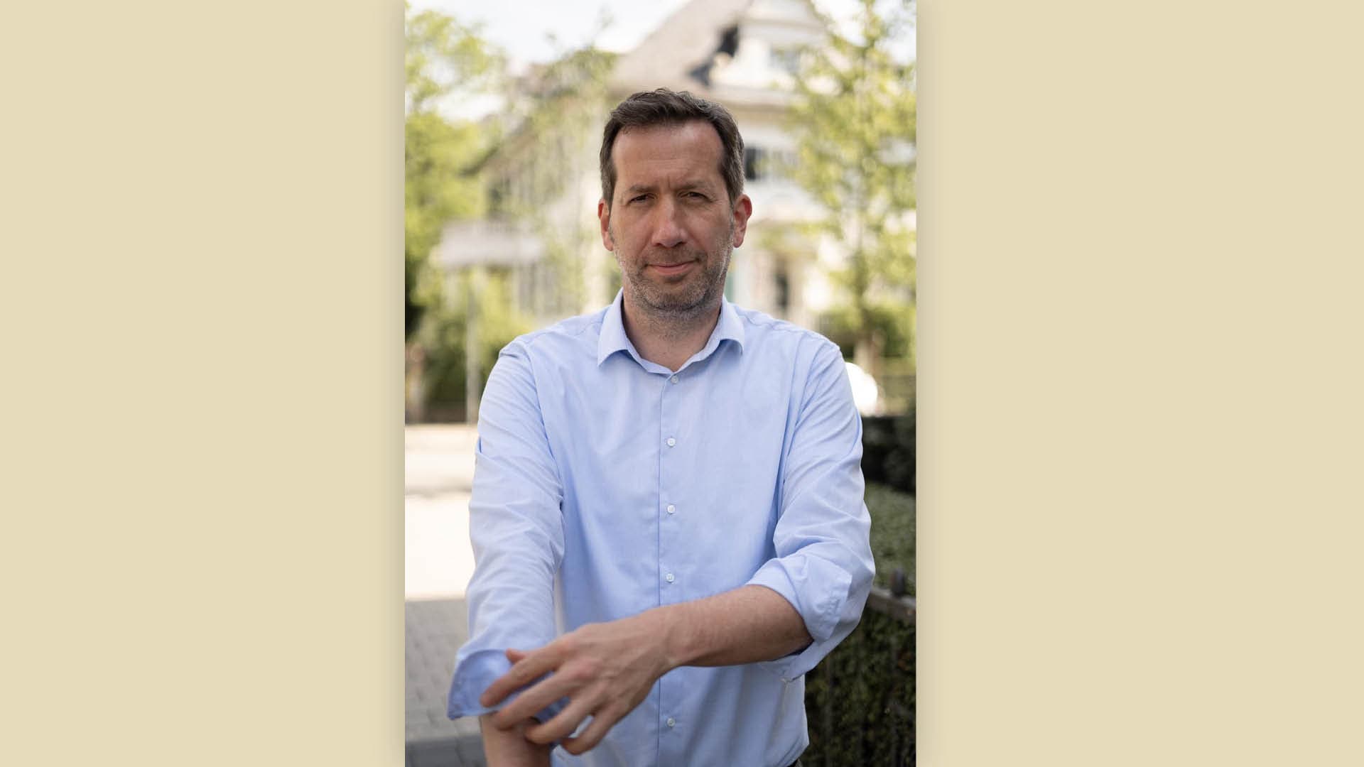 Marc Raschke im hellblauen Hemd vor unscharfem Gebäude und Grün.